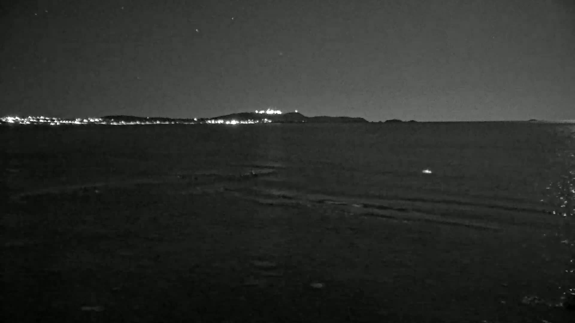 A dark, nighttime view of a calm body of water with a distant shoreline featuring lights on a hill.