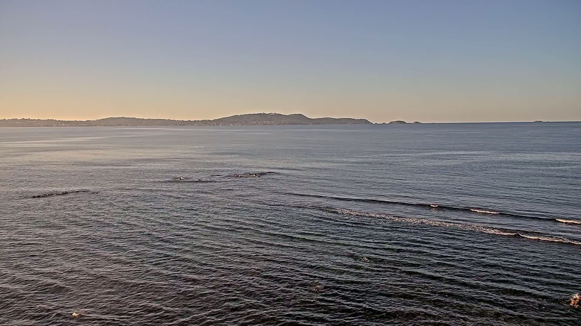 A calm ocean with small waves laps gently against the shore under a clear, light-blue sky, with a distant landmass visible on the horizon.