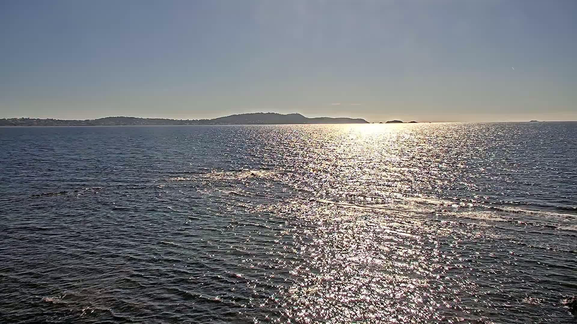 A calm, sunlit sea reflects the light, with a distant shoreline visible on the horizon under a clear sky.