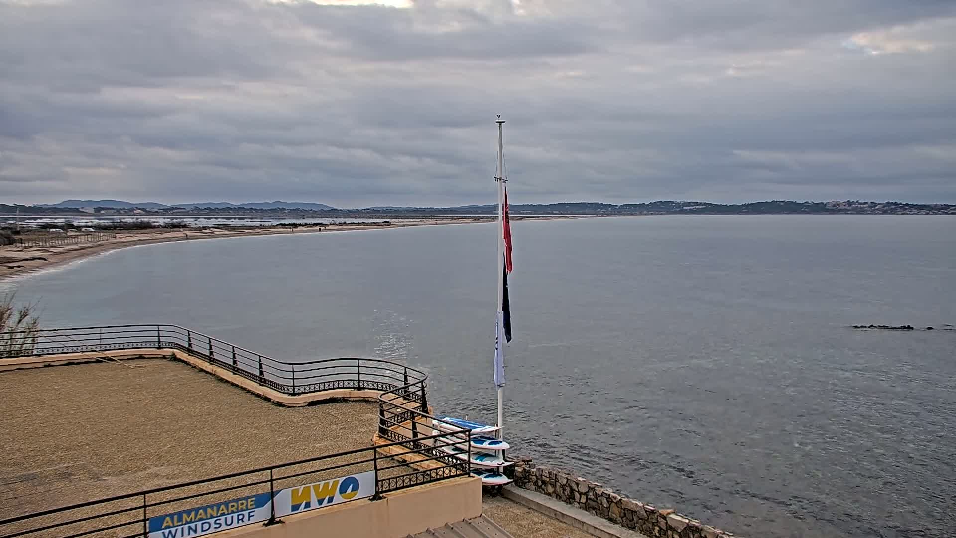 A calm, grey day shows a long sandy beach curving into a bay with calm water, a flagpole with a few flags, and a small stone structure with a gravel top and a railing.