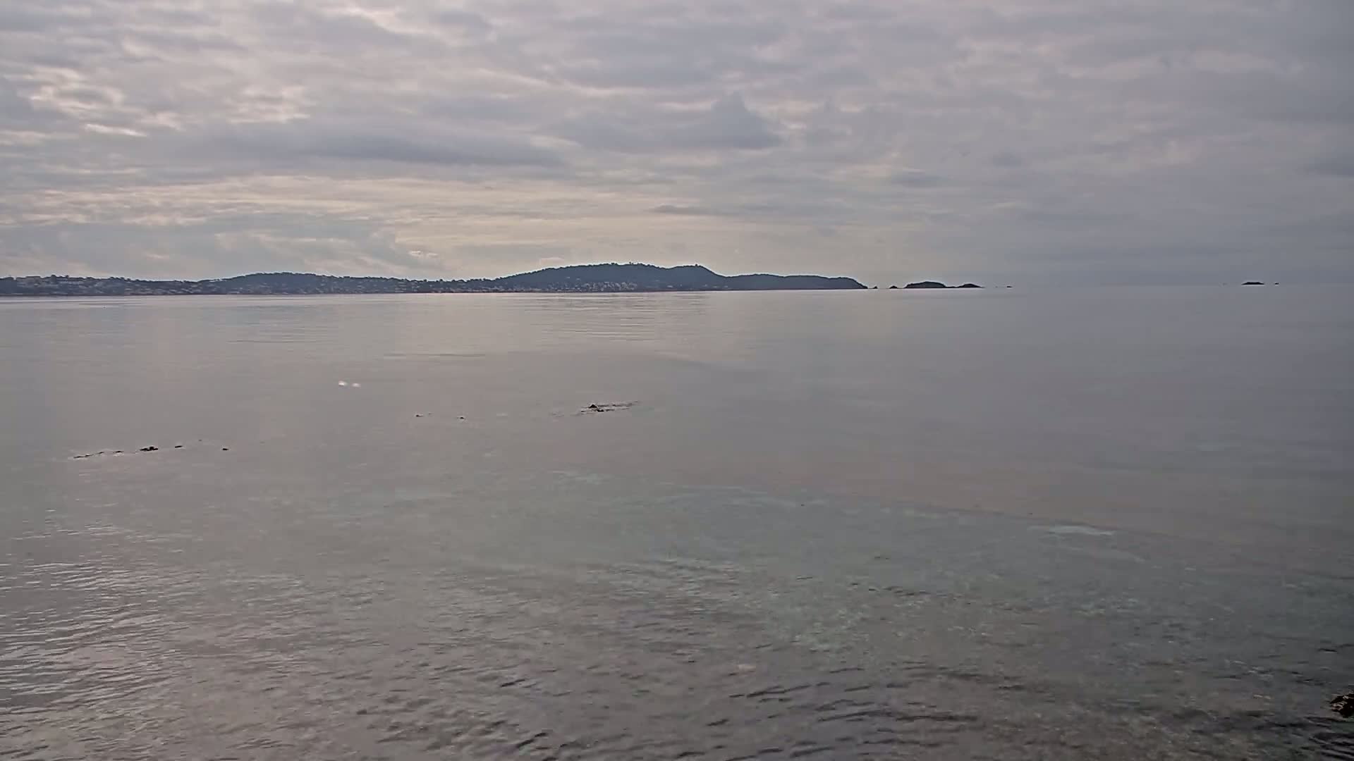 A calm, grey-sky day reveals a tranquil body of water with a distant shoreline featuring hills and small islands.