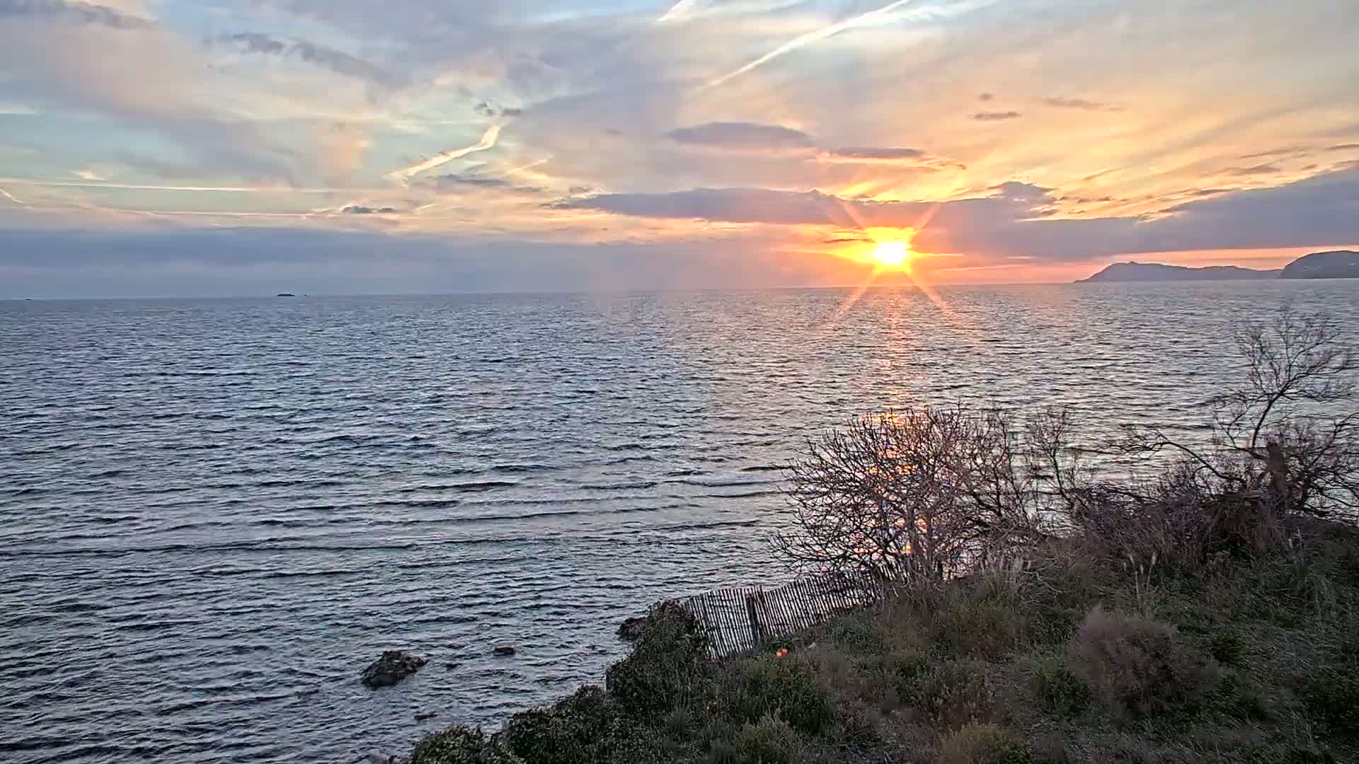 A sunset over a calm sea, with a distant landmass visible under a partly cloudy sky.