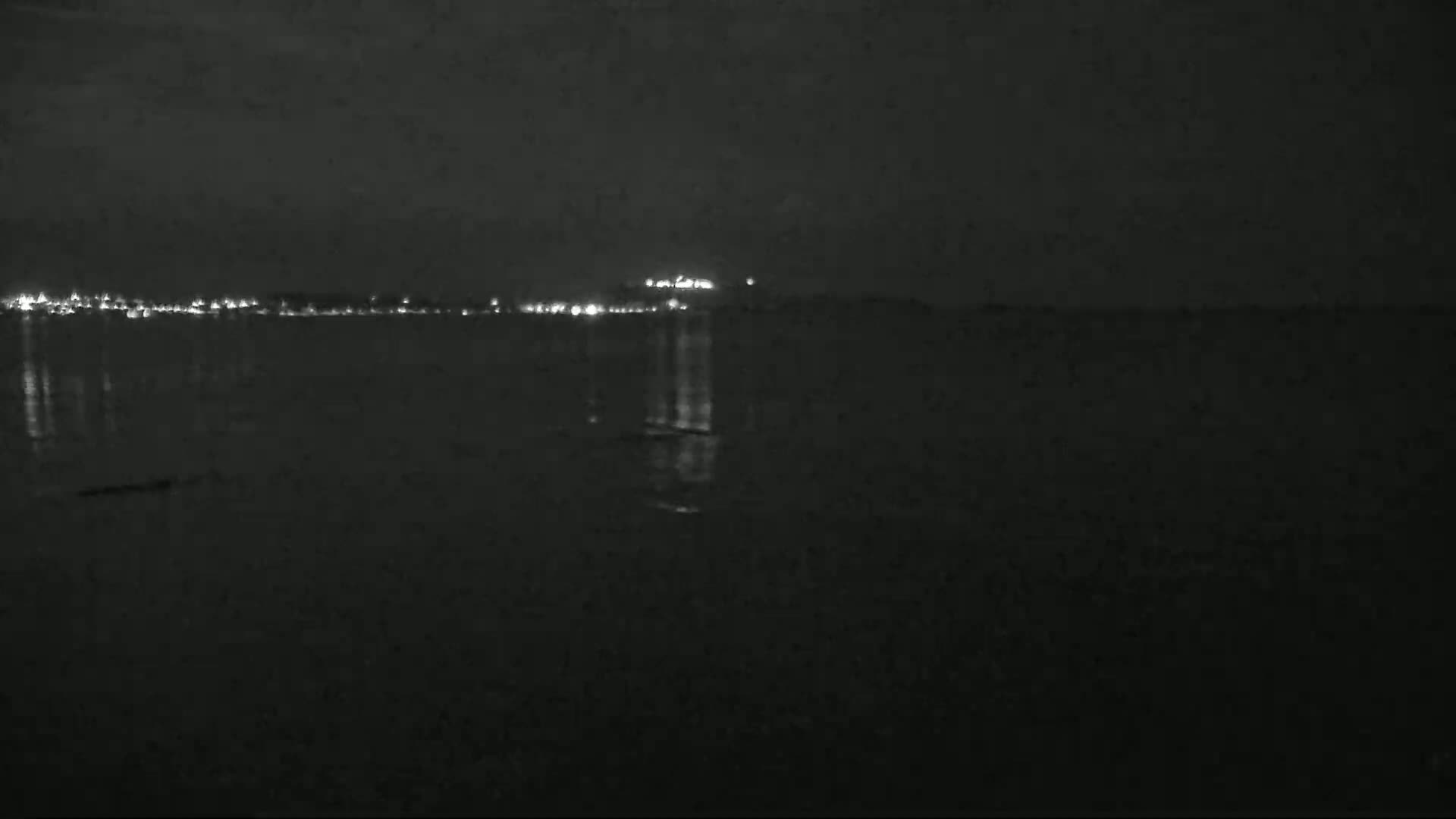 A dark nighttime scene shows a body of water reflecting the lights of a distant city under a clear sky.