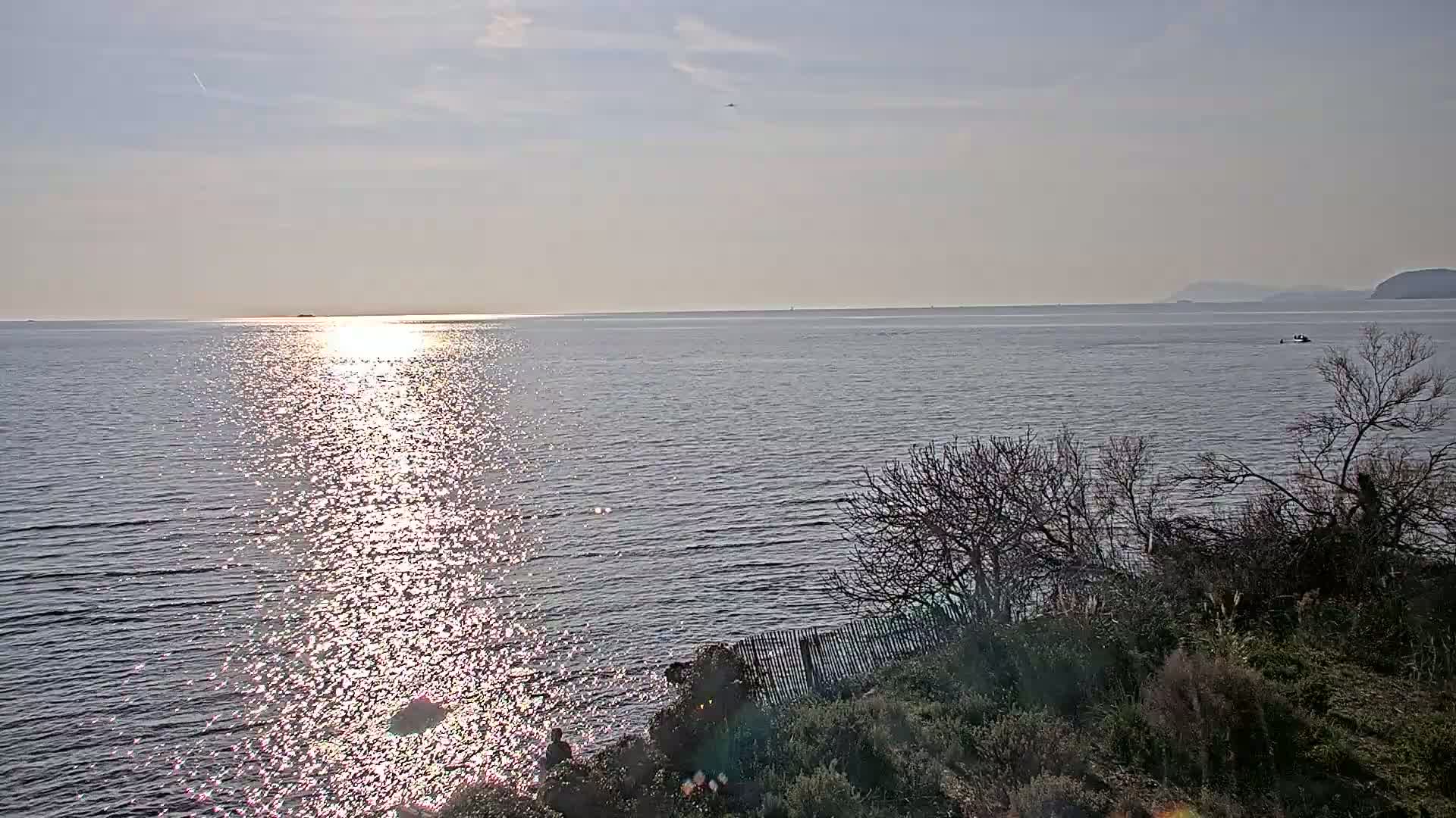 A calm sea reflecting sunlight shimmers under a mostly clear sky, viewed from a rocky shoreline with sparse vegetation.