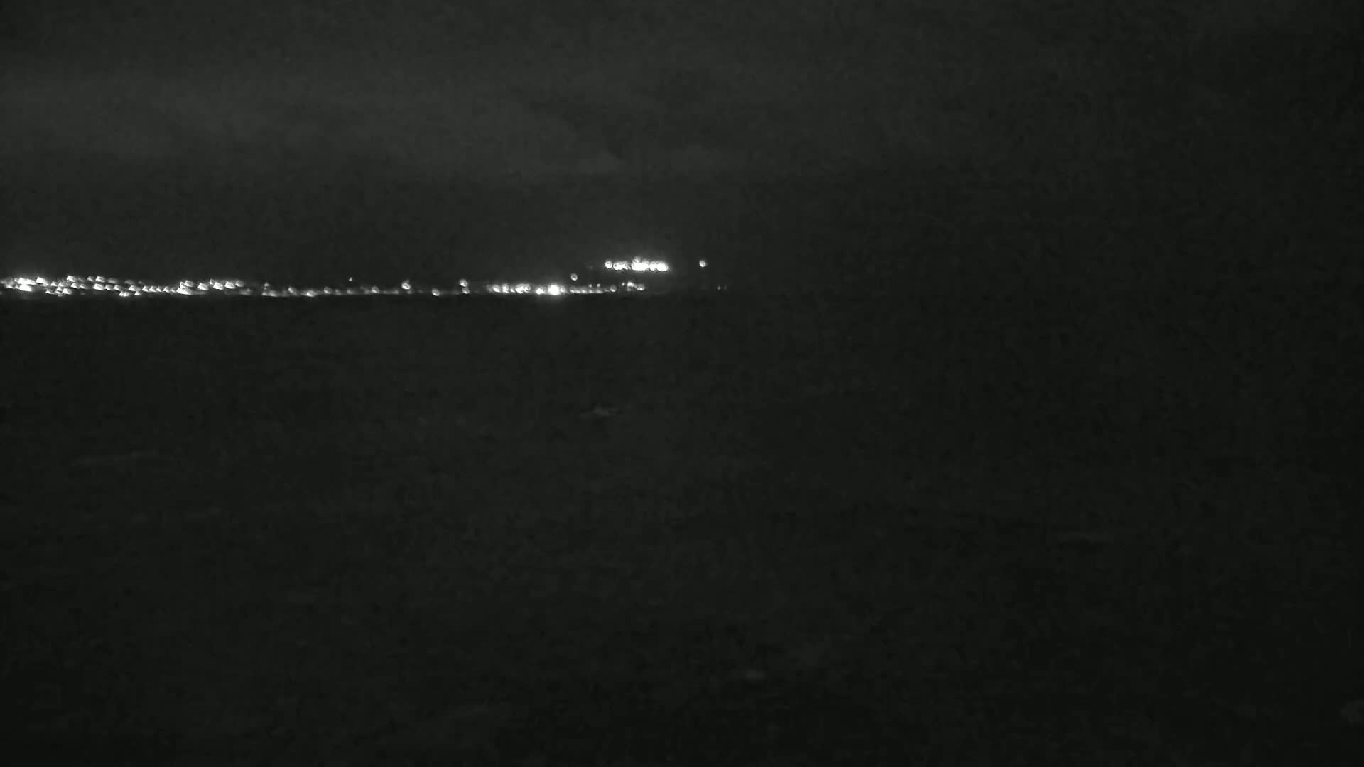 A dark, nighttime scene shows a distant line of lights across a dark landscape under a cloudy sky.