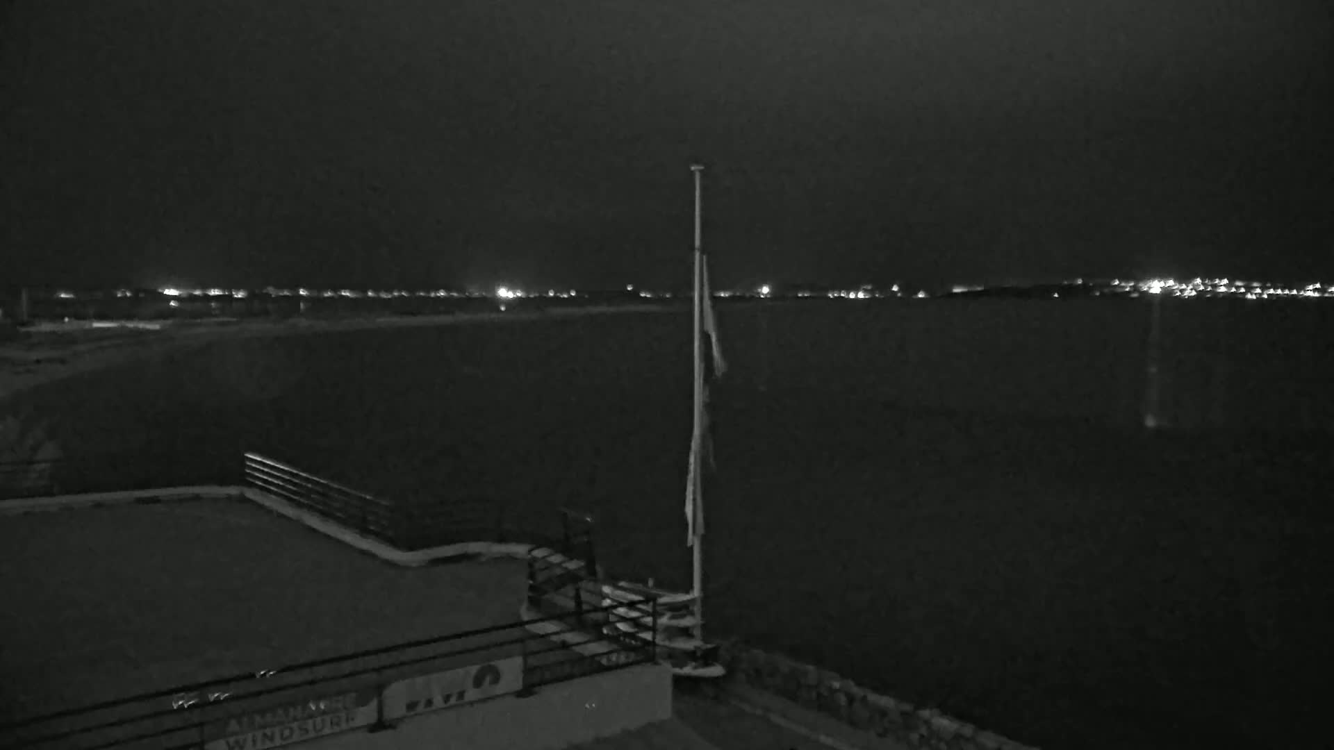 A nighttime view of a dark body of water with a distant line of lights, a flagpole with a tattered flag, and a railing on a structure in the foreground.