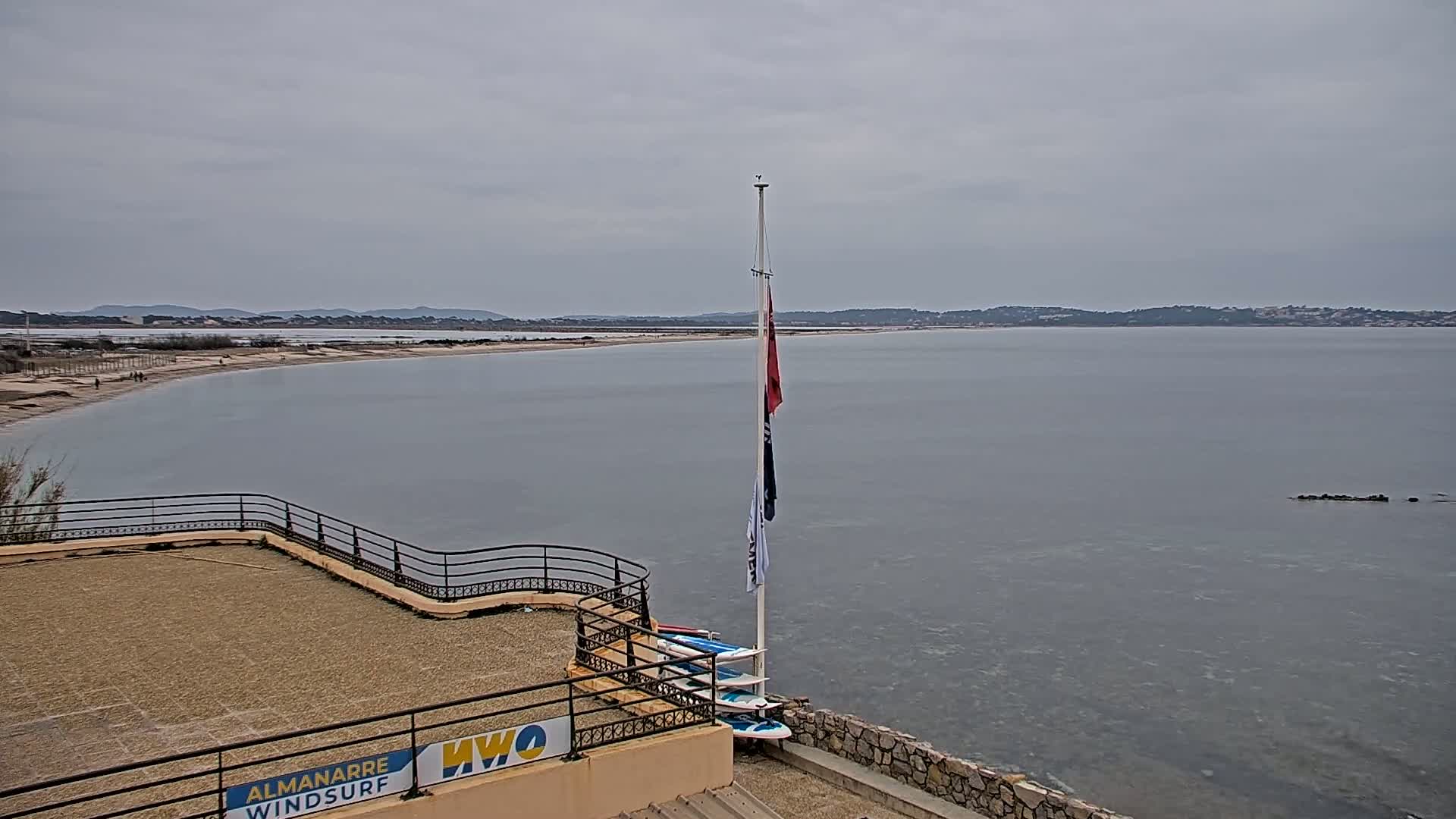 A calm, grey day overlooks a sandy beach and calm body of water, with several windsurfing boards leaning against a building.