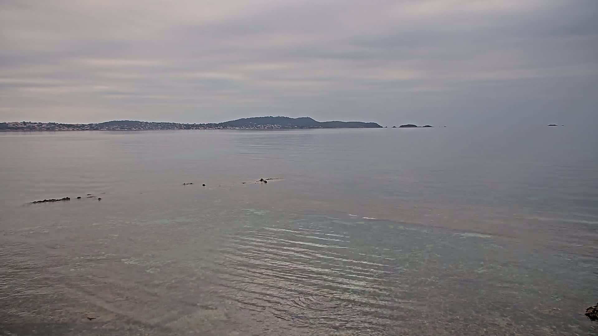 A calm, overcast day reveals a shoreline with shallow, clear water, and a distant landmass with low hills.