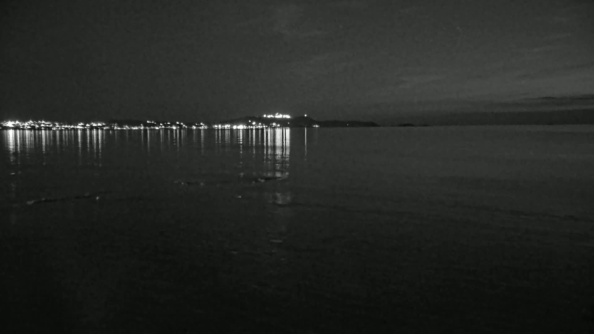 A nighttime view of a calm body of water reflecting the lights of a town on a distant hill.