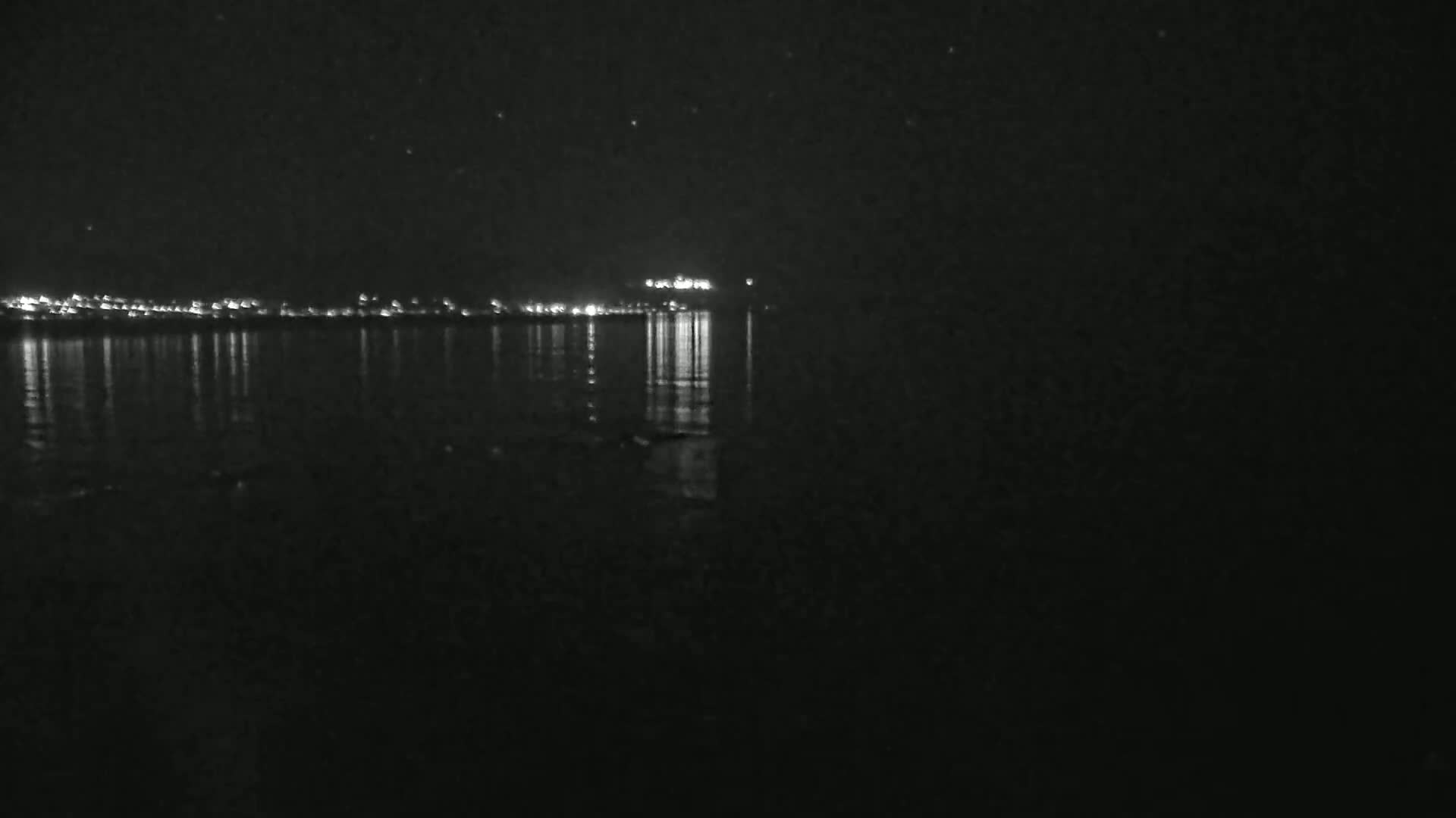 A nighttime view of a calm body of water reflecting the lights of a distant town under a clear, starry sky.