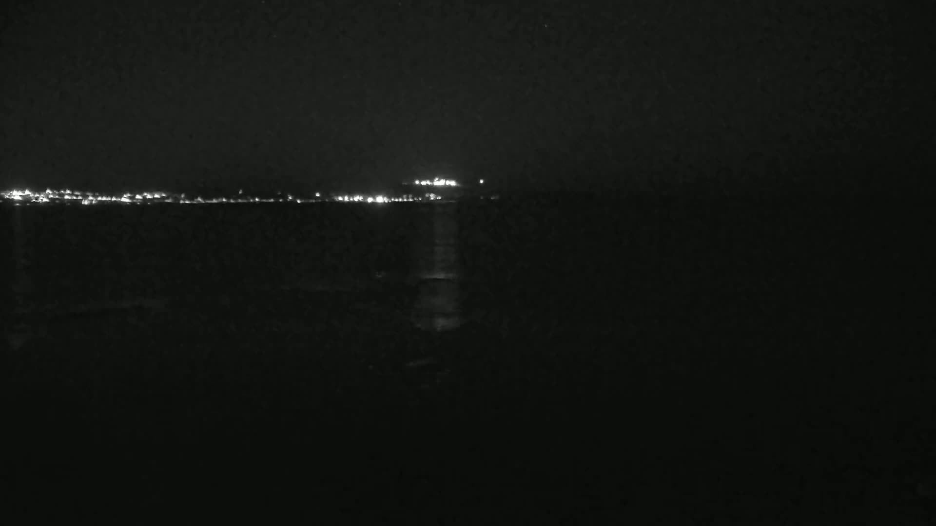 A dark, nighttime scene shows a distant shoreline illuminated by lights reflecting on dark water.