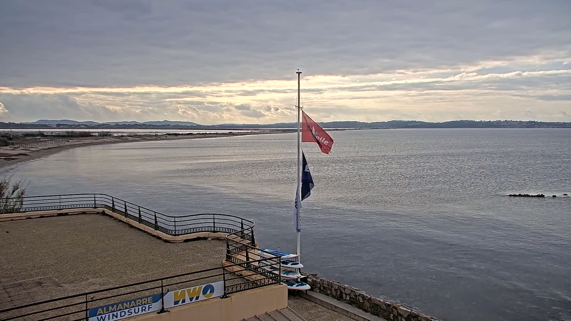 A calm body of water stretches to a distant shoreline under a partly cloudy sky, with a flagpole bearing several flags standing on a paved area next to a railing overlooking the water.