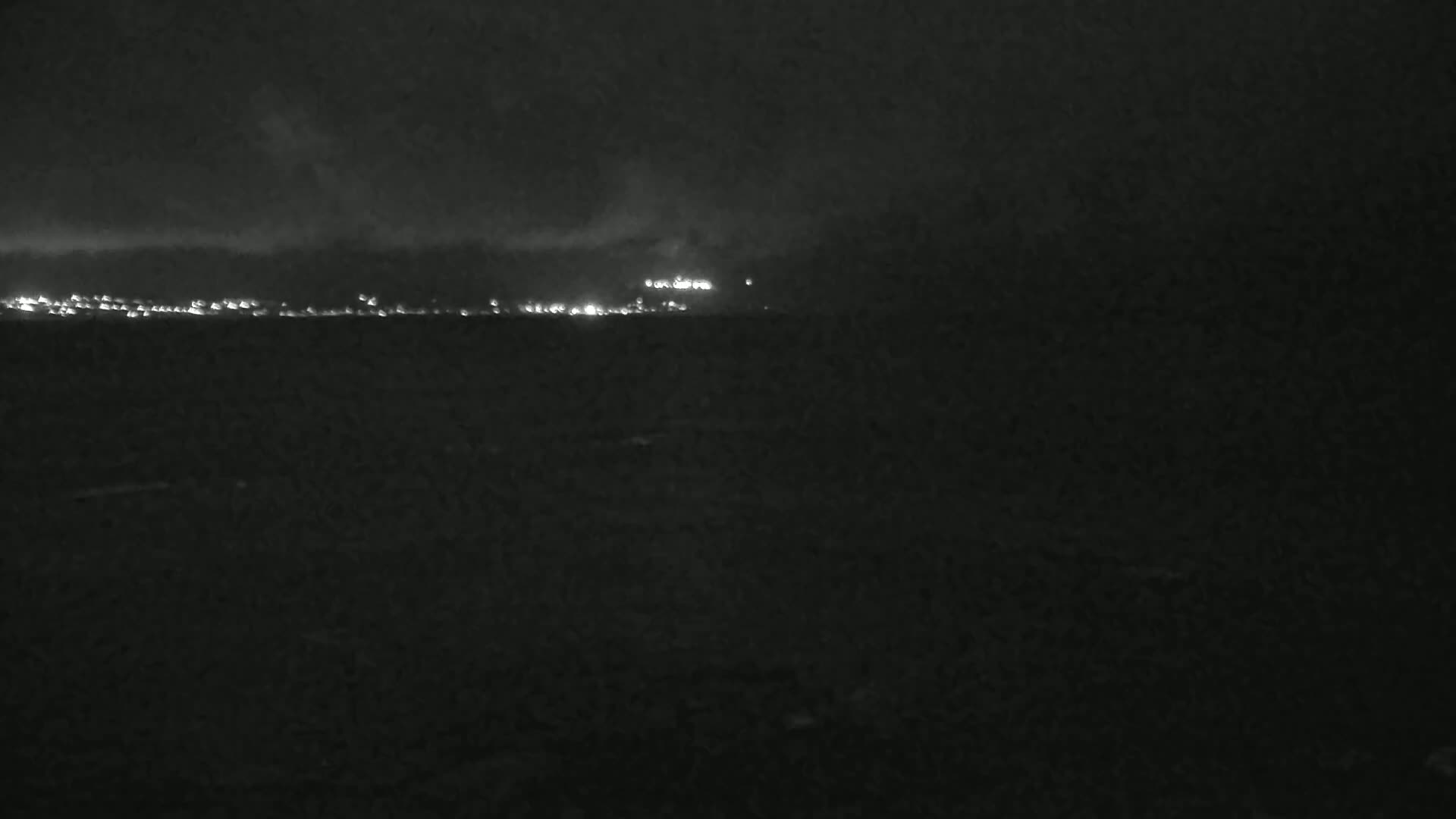 A nighttime view shows a distant line of lights across a dark body of water under a cloudy sky.
