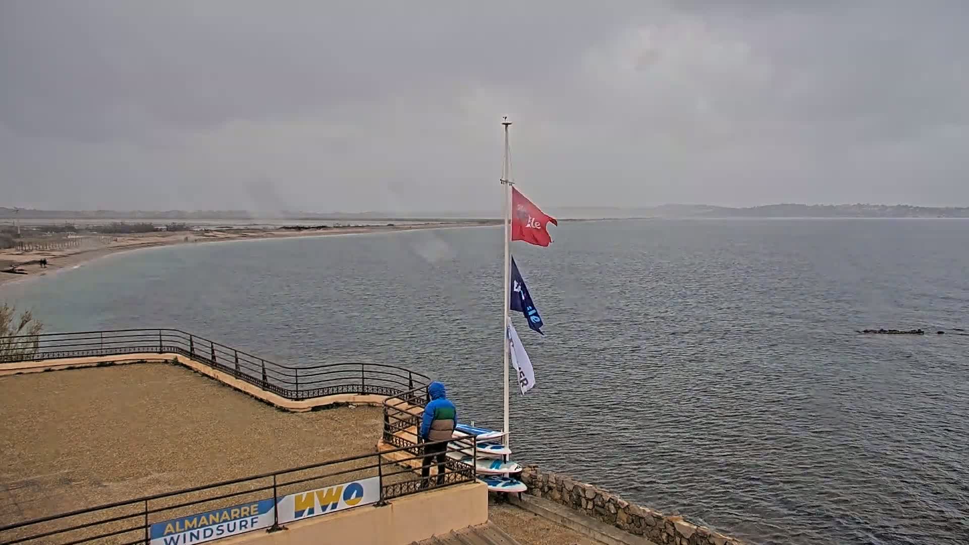A person stands near several windsurf boards on a raised platform overlooking a calm body of water on an overcast day.