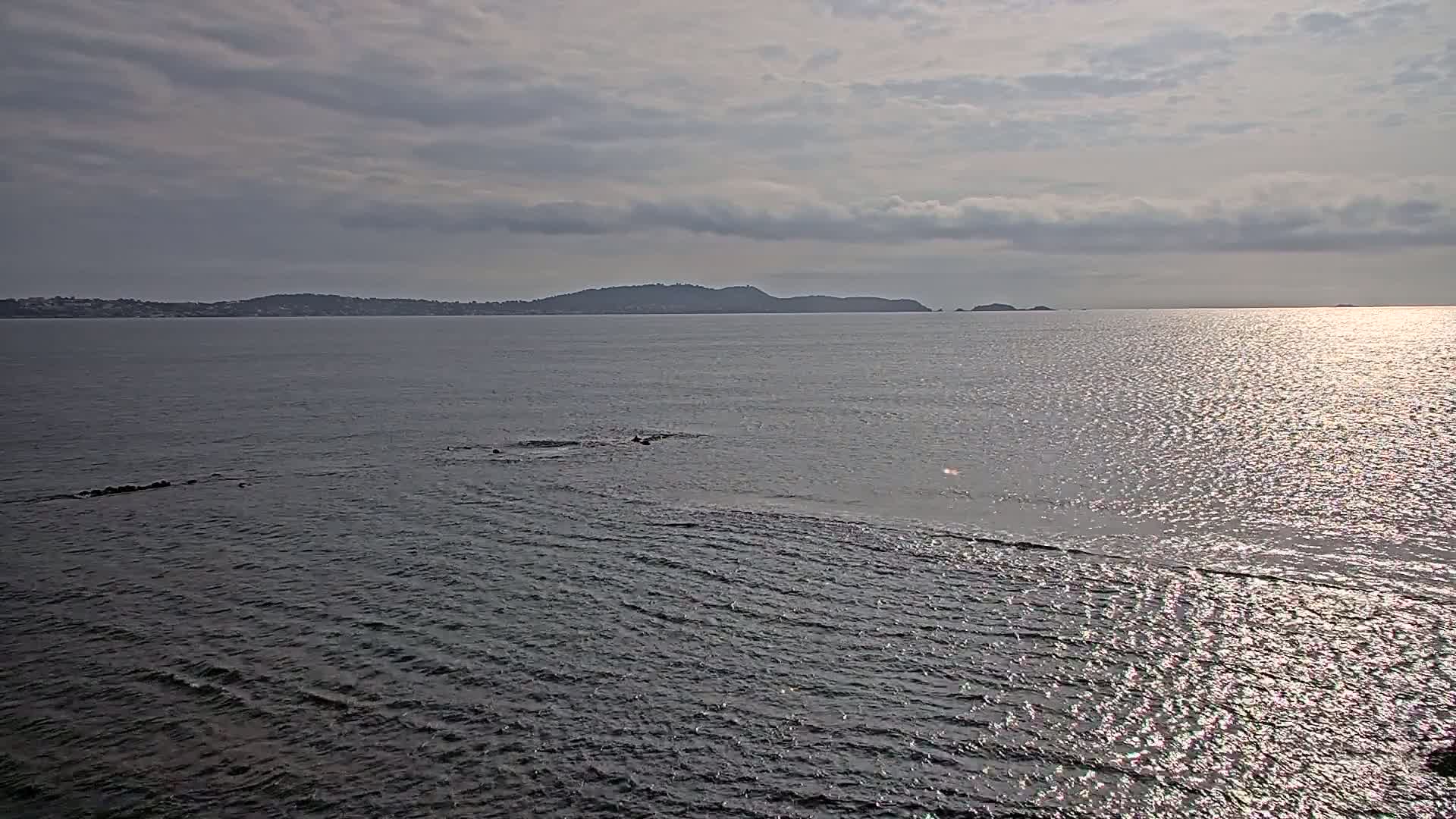 A calm sea with small waves reflects sunlight under a cloudy sky, showing a distant shoreline with hills in the background.