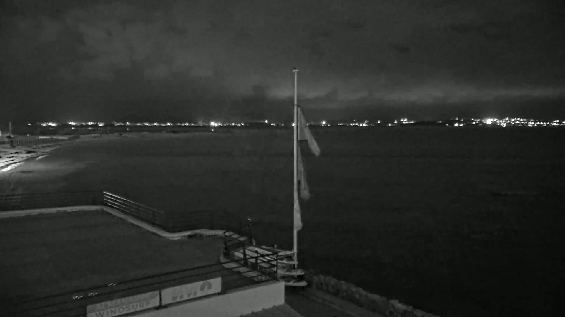 A nighttime view of a dark beach and body of water, with a distant city skyline illuminated, under a cloudy sky.