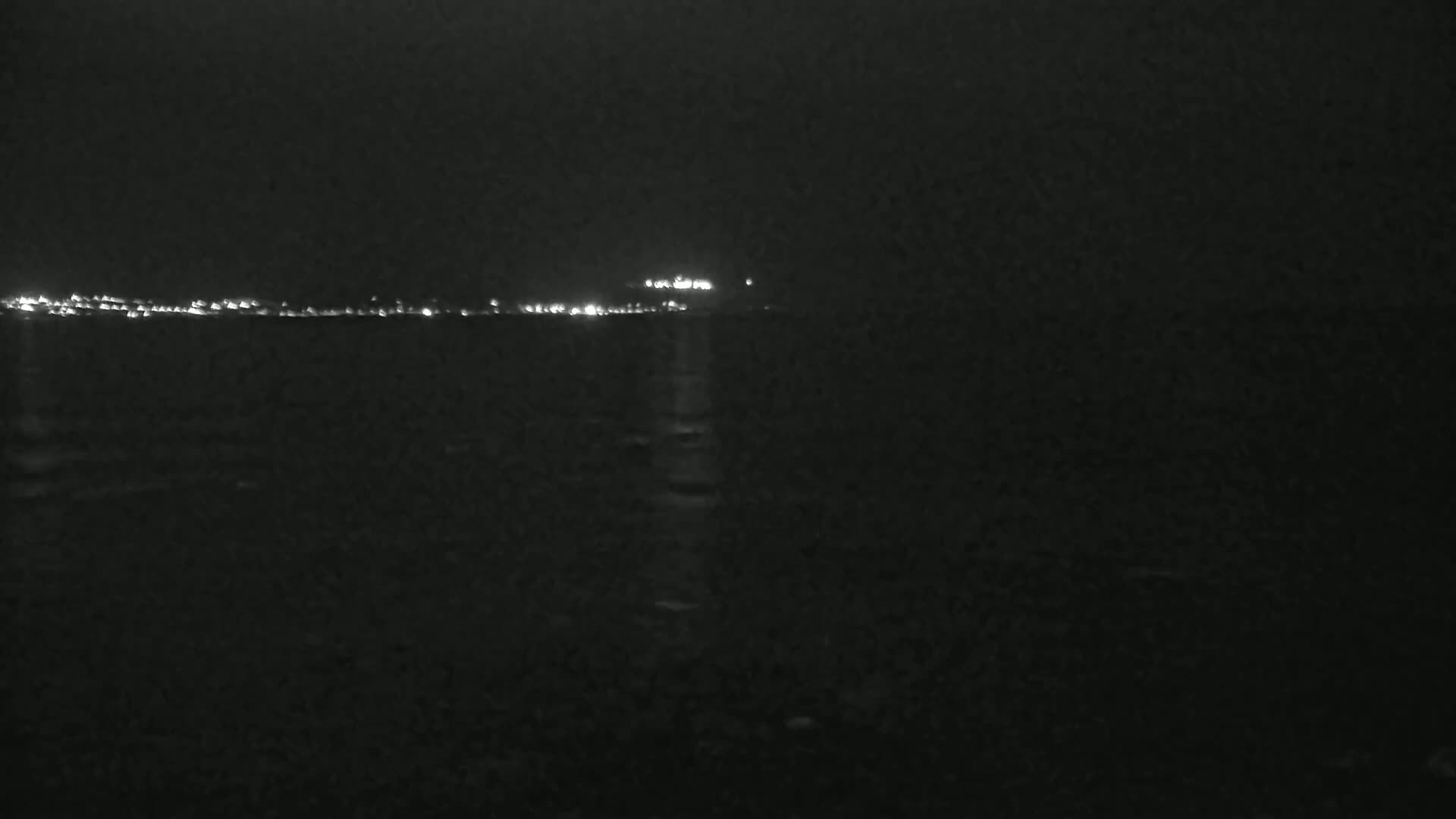A dark, nighttime scene shows a distant, faintly lit shoreline across a body of dark water.