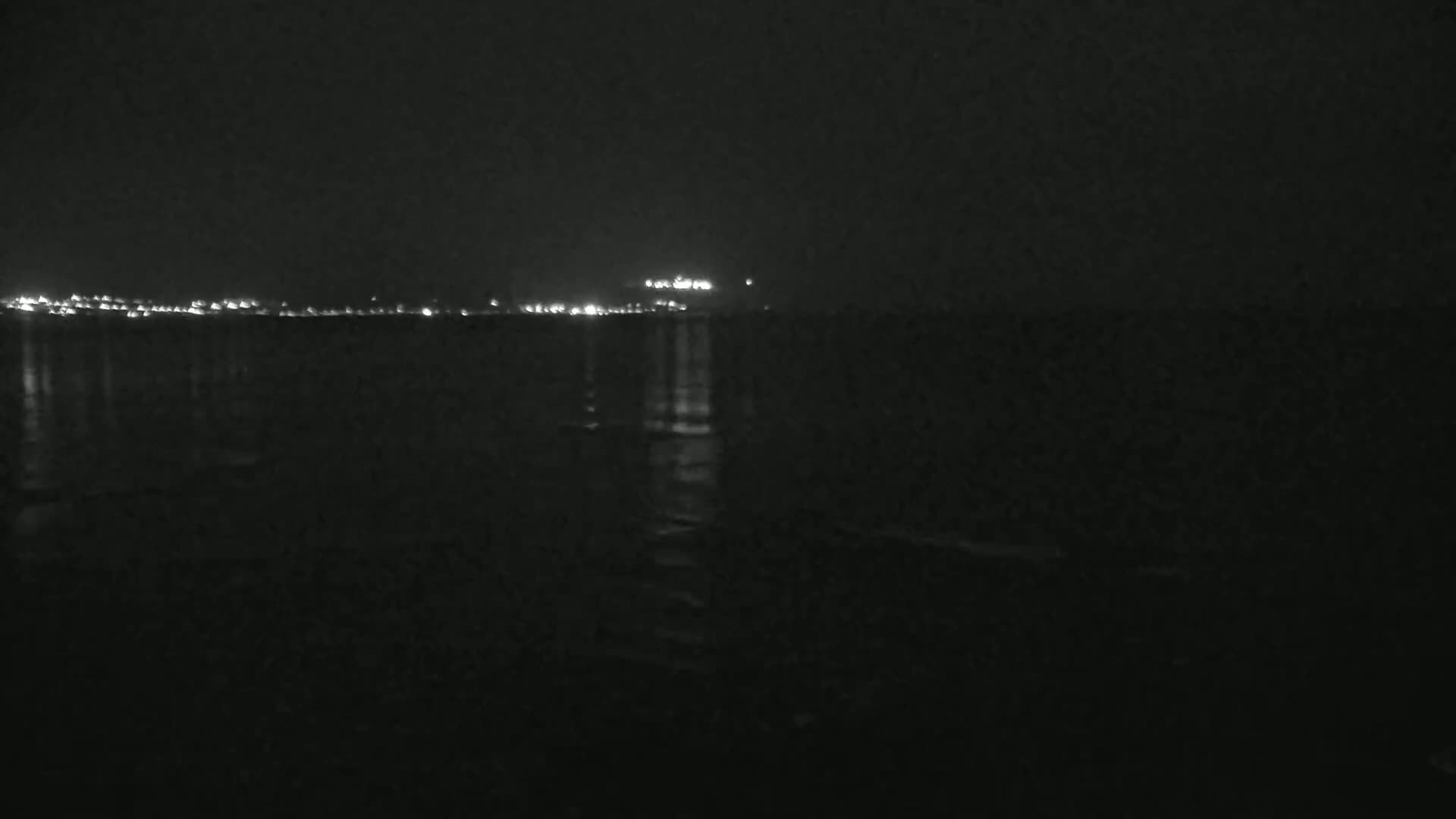 A dark, nighttime scene shows a calm body of water reflecting the lights of a distant city under a clear sky.
