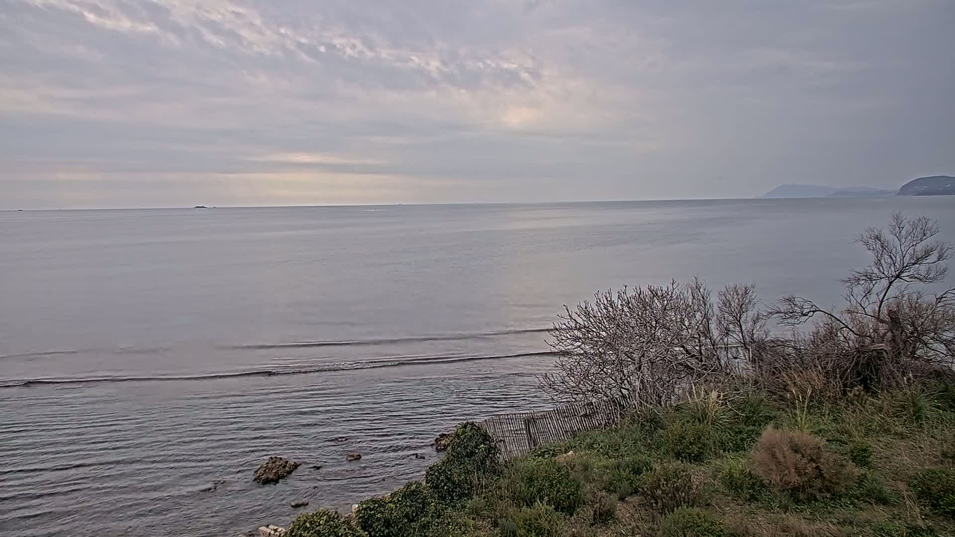 A calm sea with small waves laps a rocky shoreline under a cloudy sky, partially obscuring a distant mountain range.
