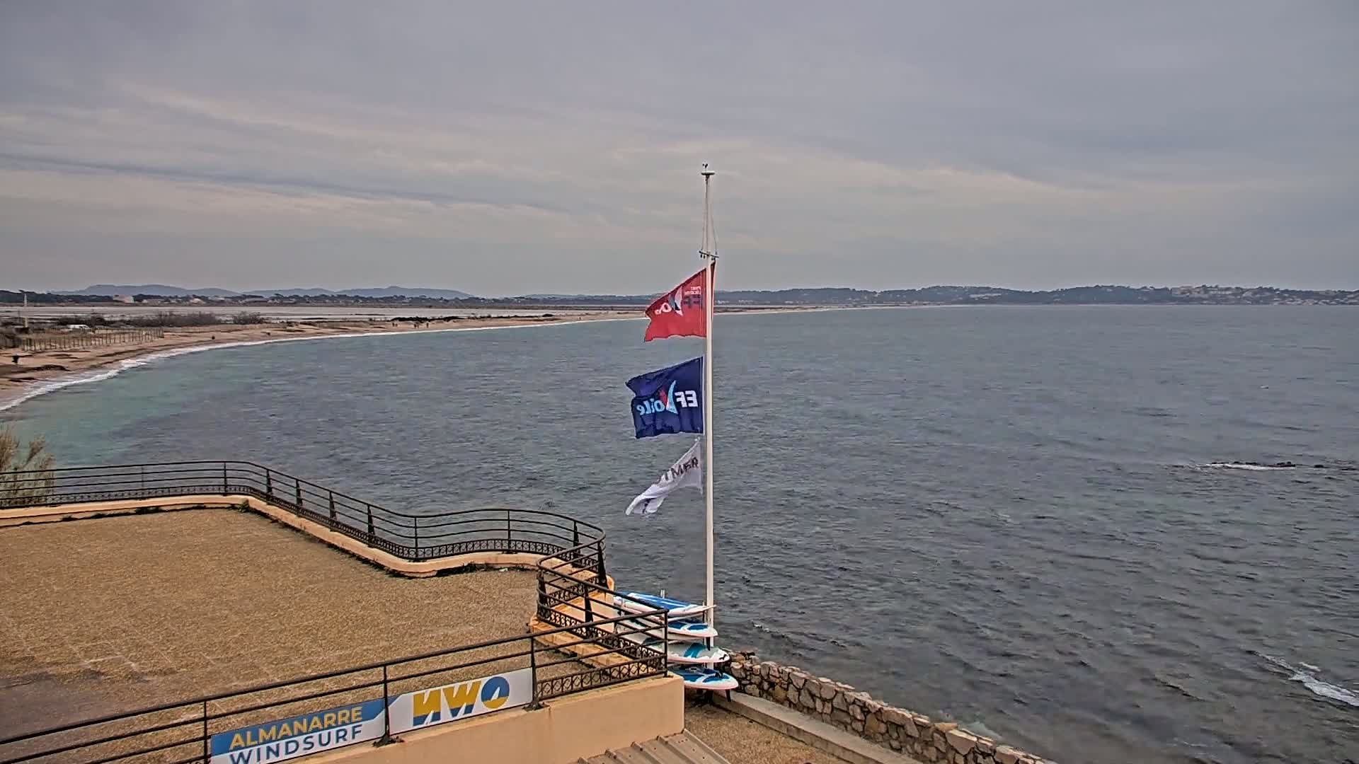 A sandy beach and calm ocean are visible under an overcast sky, with a flagpole displaying several flags and windsurf boards leaning against it on a raised platform.