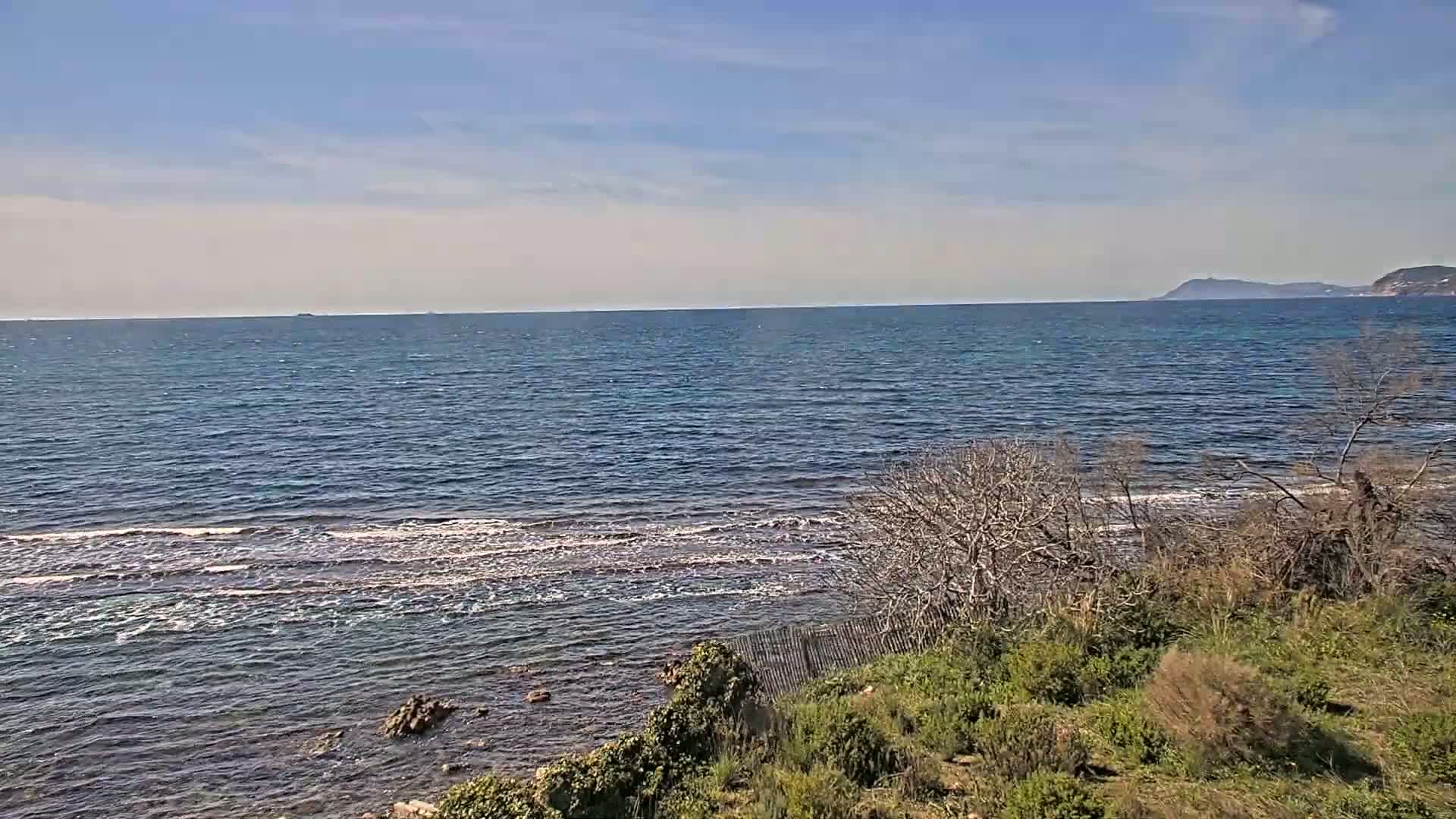 A calm sea with small waves laps a rocky shore under a mostly clear, bright sky, where sparse vegetation grows on the land.
