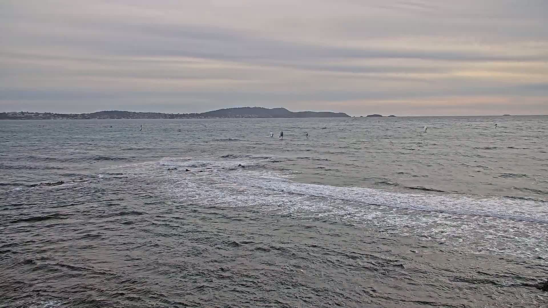 A calm, overcast day shows a body of water with several windsurfers and a distant shoreline.