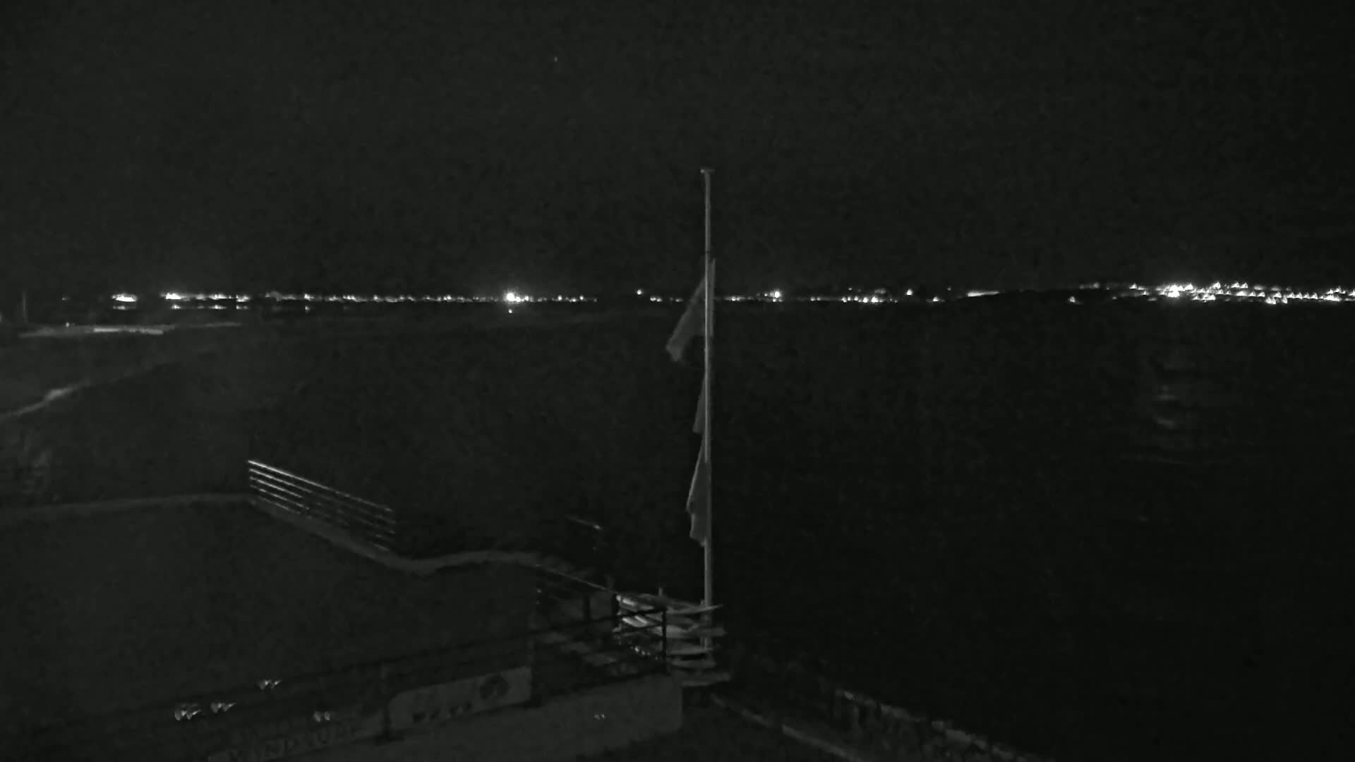 A nighttime view shows a flagpole with flags hanging limply, a railing, and a distant line of city lights under a dark sky.