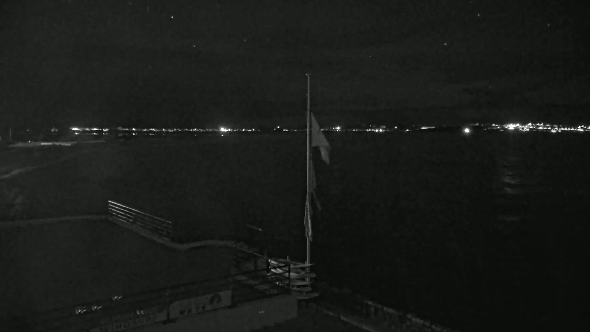 A nighttime view shows a flagpole with a partially visible flag near a railing overlooking a dark body of water with a distant city skyline illuminated by lights.