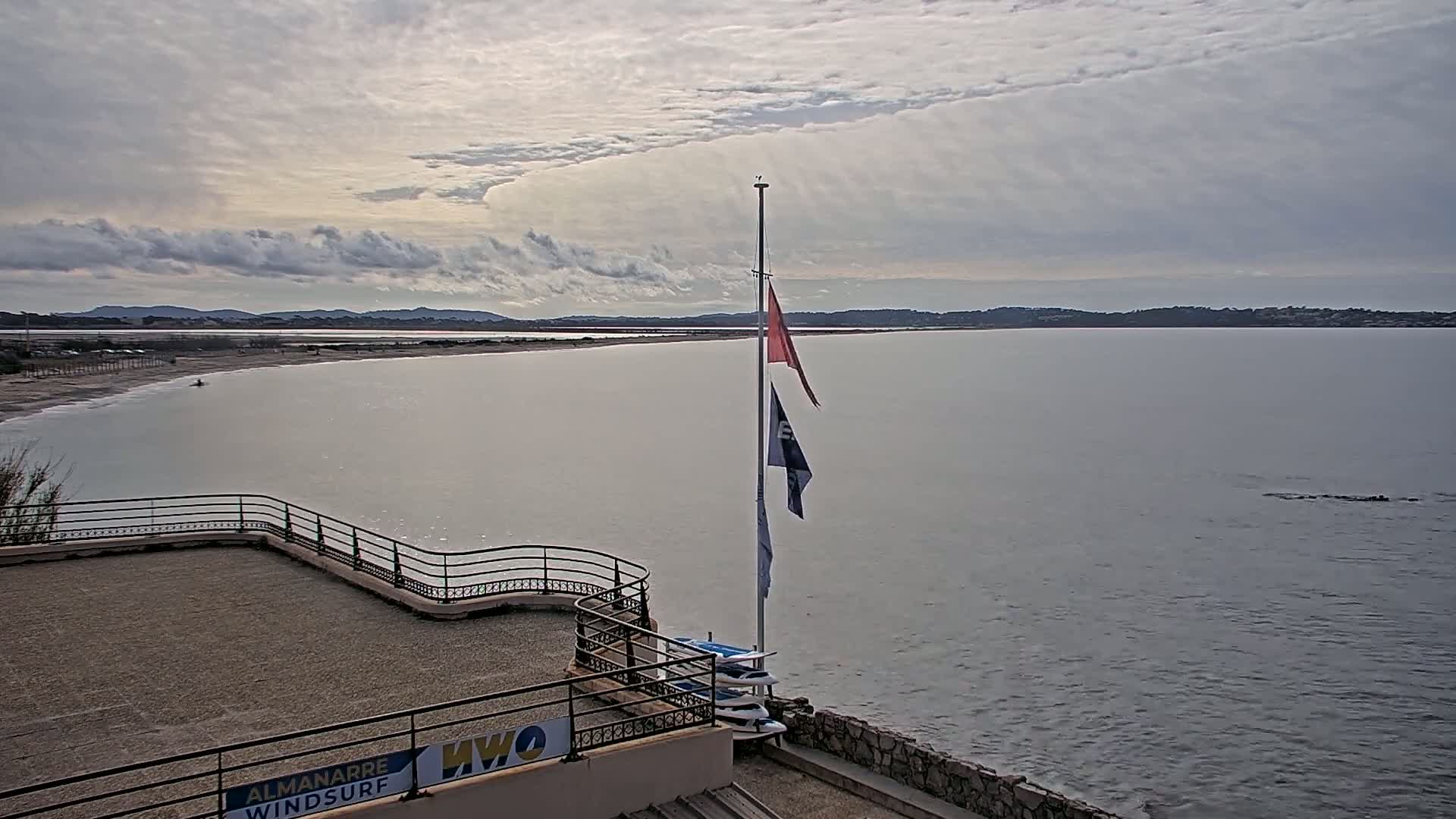 A calm, overcast day reveals a view of a beach, a body of water, and distant land under a cloudy sky; several windsurfing boards are stacked near a flagpole displaying two flags.