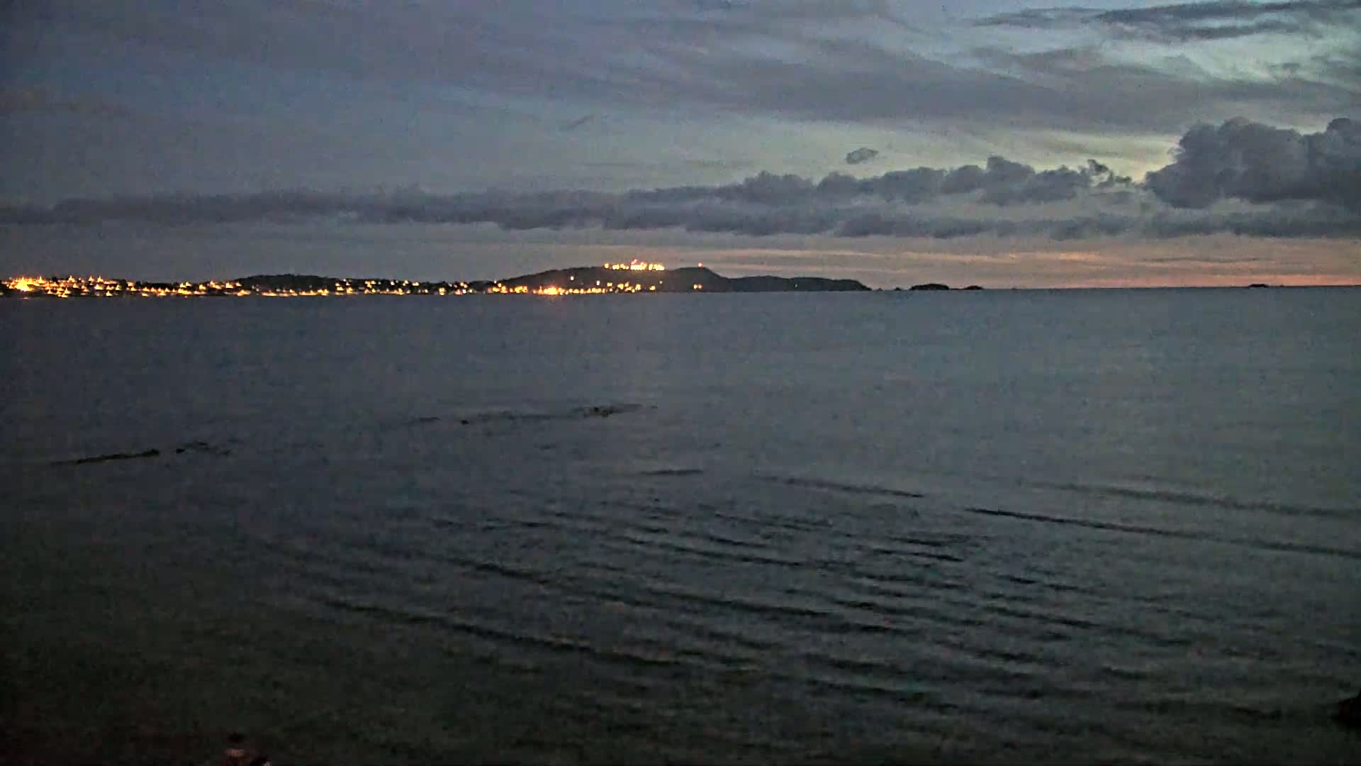 A dark, calm sea stretches toward a distant, faintly lit coastline under a mostly cloudy, twilight sky.