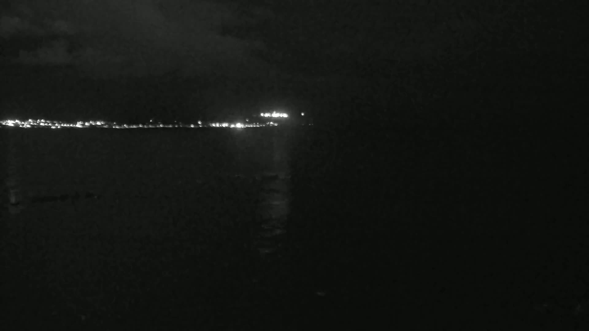 A dark, nighttime scene shows a distant line of lights on the horizon over a dark body of water under a cloudy sky.