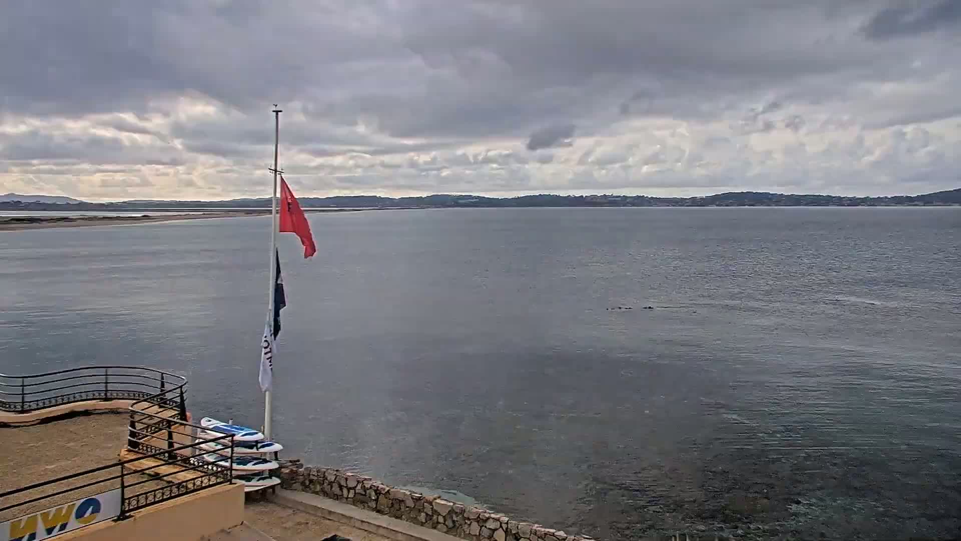 A calm body of water stretches to a distant shoreline under a cloudy sky, with a flagpole displaying a red and white flag and several surfboards leaning against a low wall.