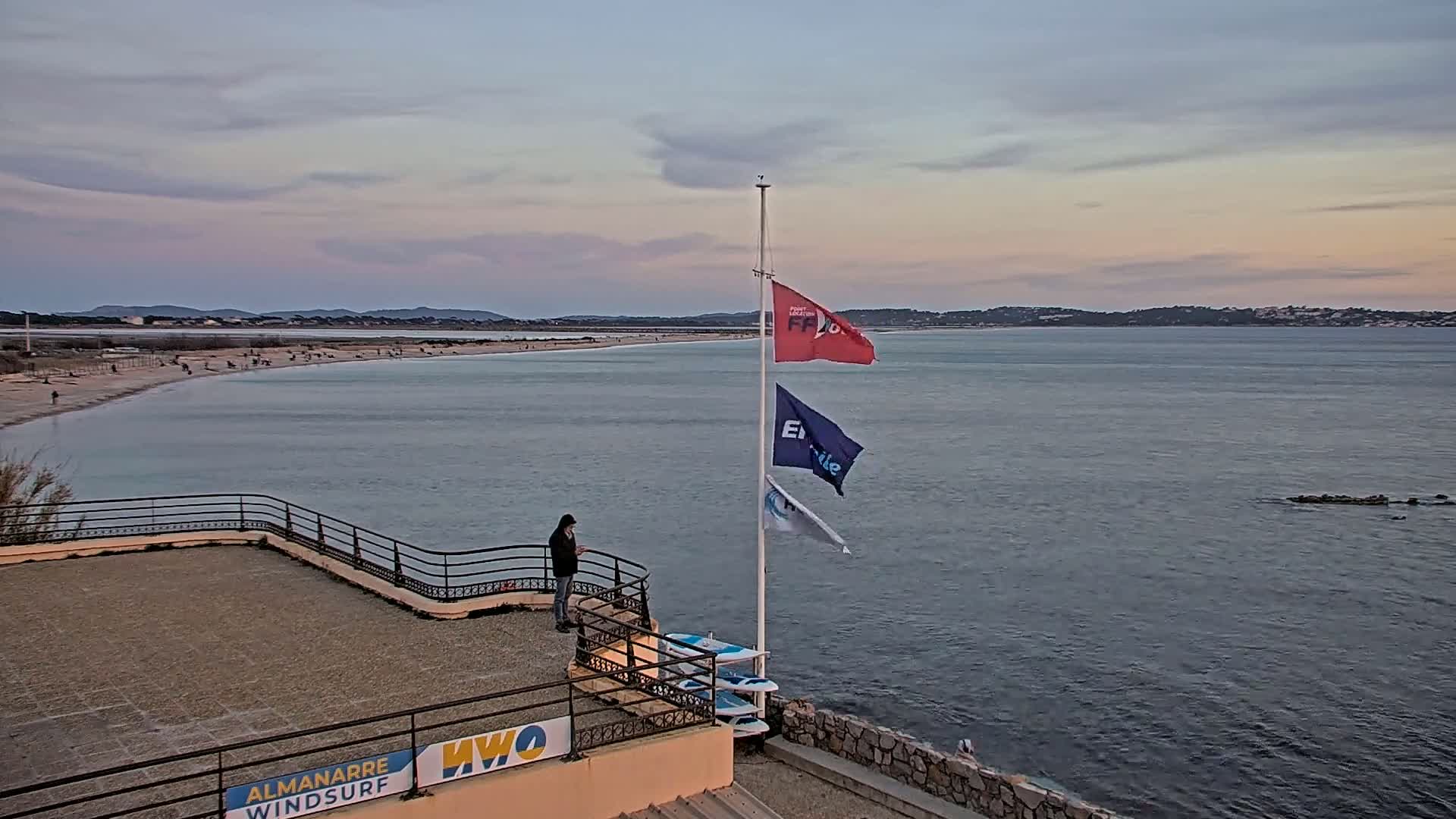 A person stands on a rooftop overlooking a calm bay with a sandy beach and distant shoreline under a partly cloudy, dusky sky.
