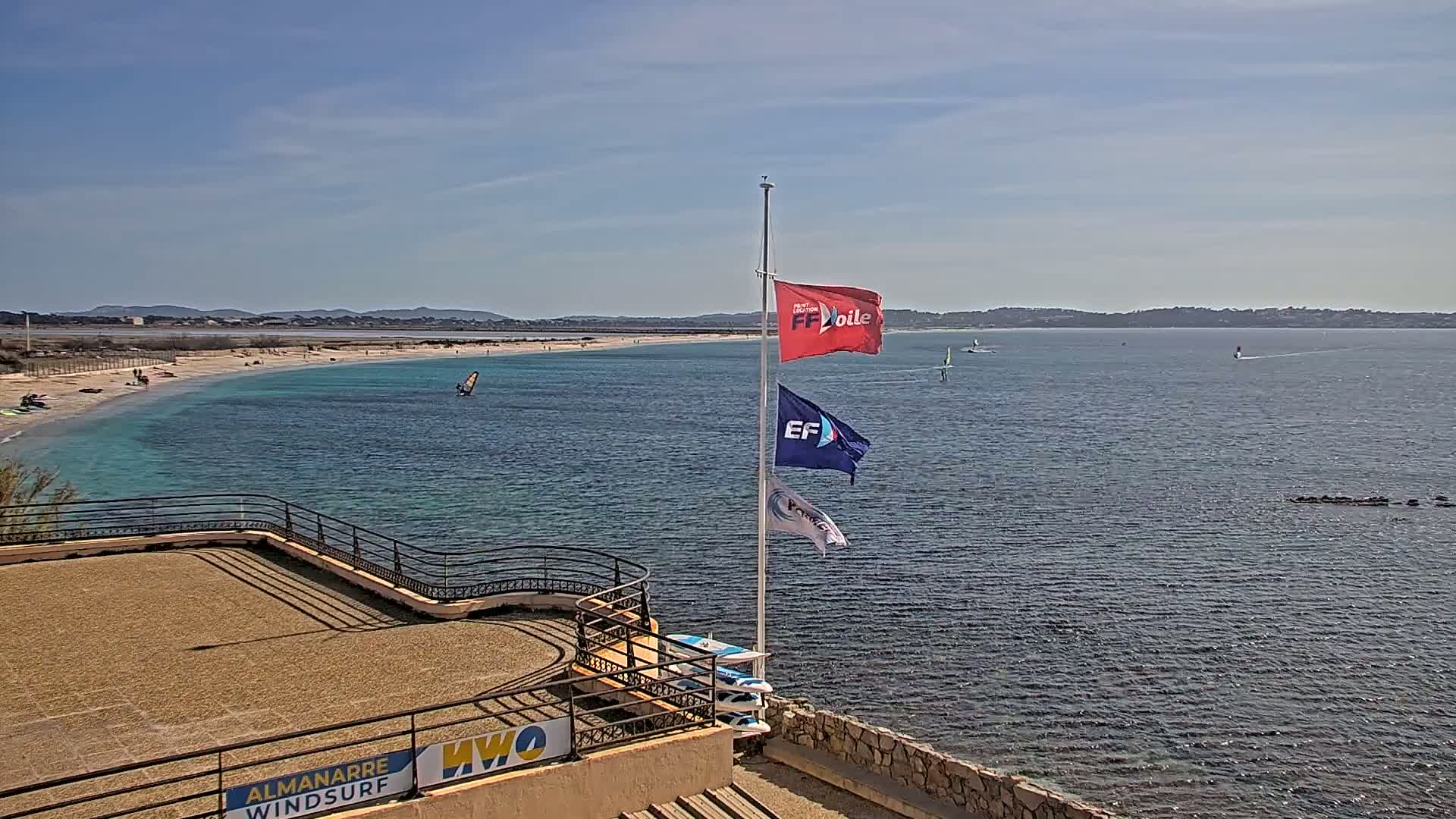 An elevated view of a sandy beach and calm, turquoise water with windsurfers, under a clear, sunny sky.