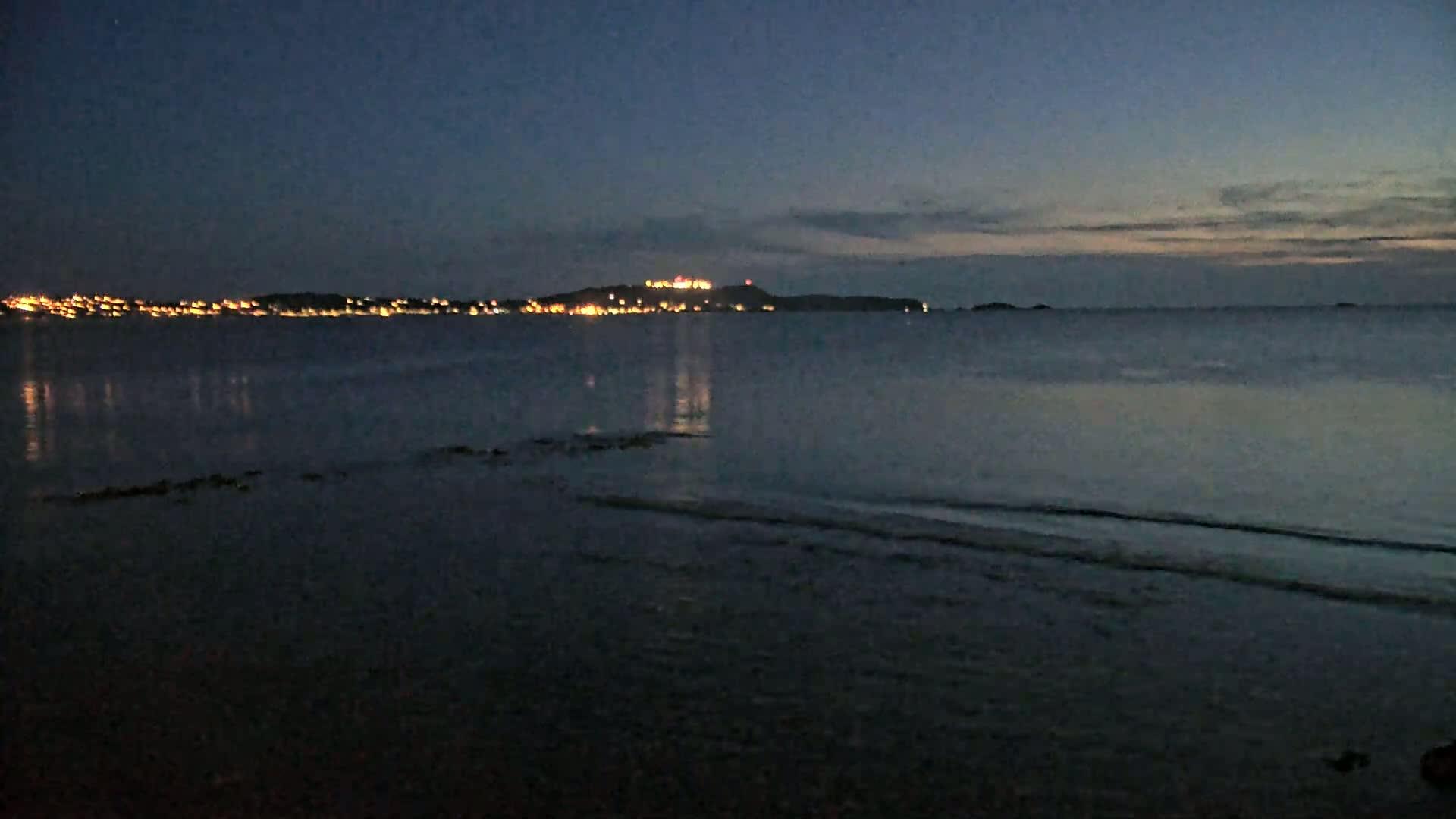 A dark, calm body of water reflects the lights of a distant town on a hill under a twilight sky.