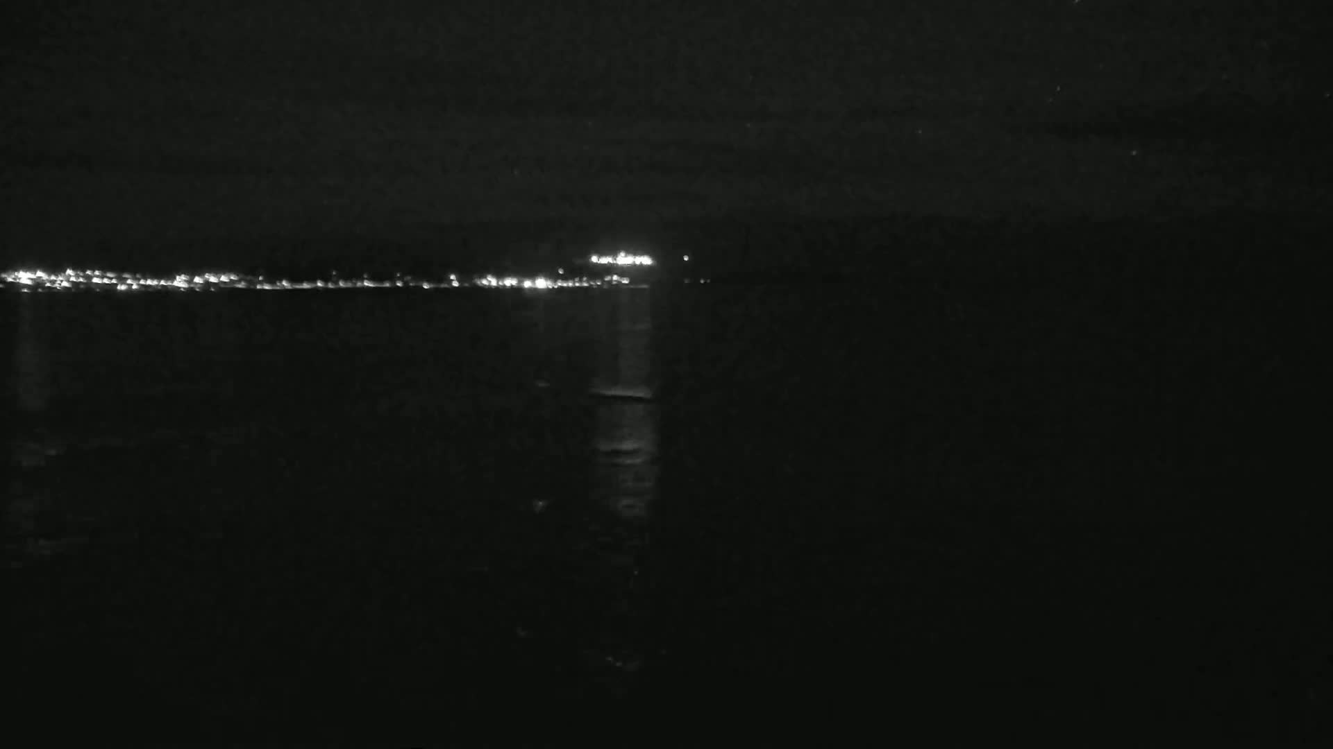 A nighttime view of a dark body of water reflecting the lights of a distant city under a clear, dark sky.