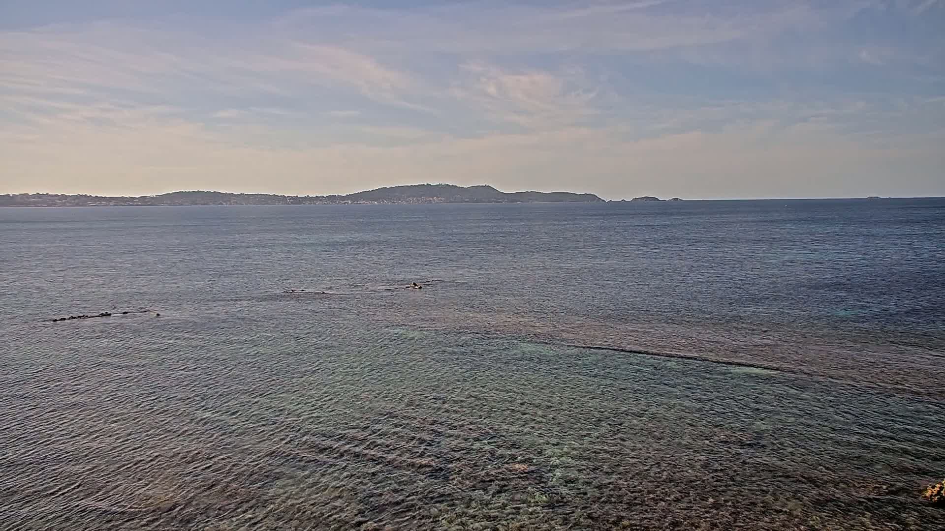 A calm, clear sea stretches to a distant island under a mostly cloudy sky.