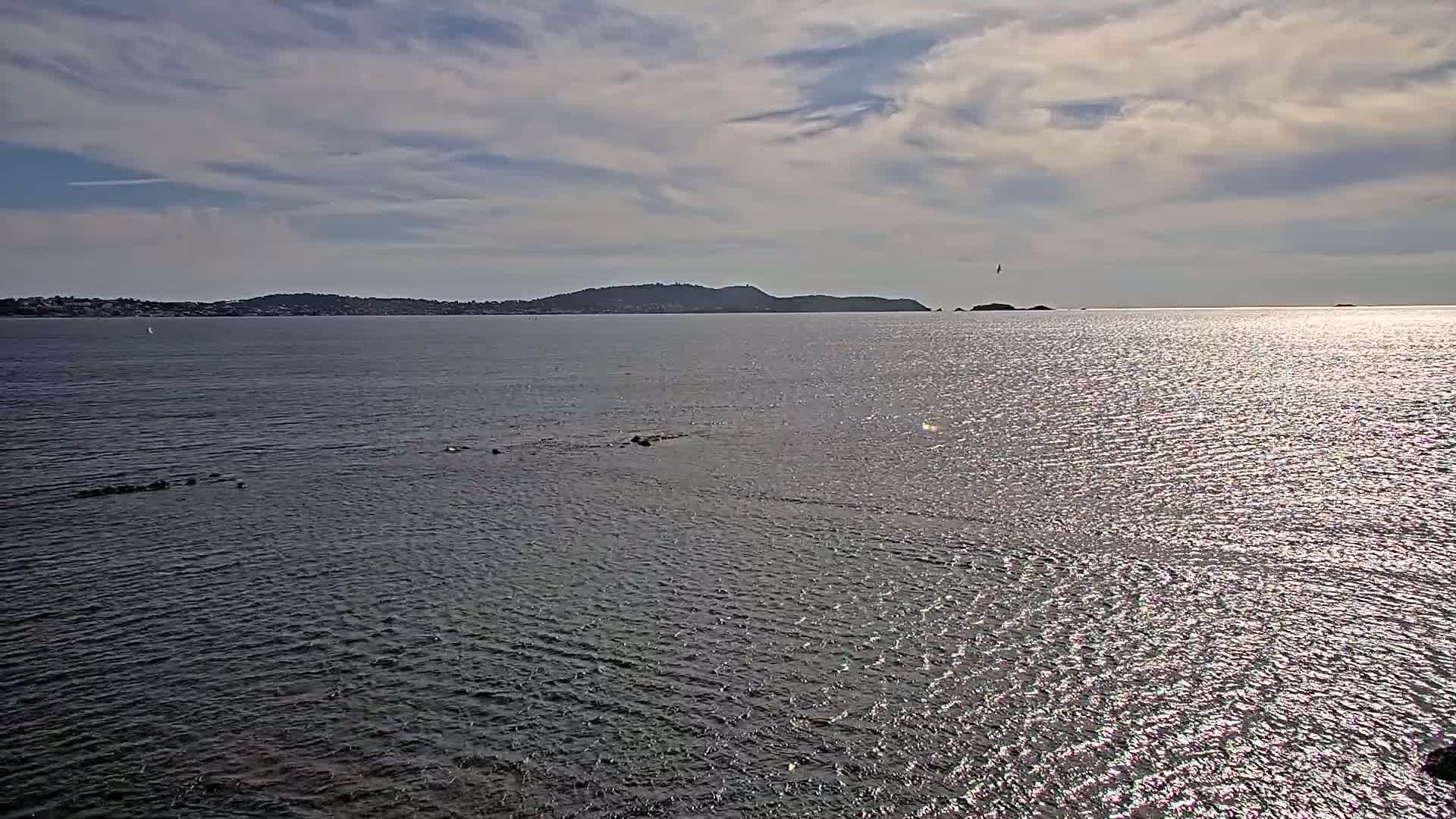 A calm, sunlit body of water stretches to a distant shoreline featuring a low-lying, dark-green hill under a partly cloudy sky.