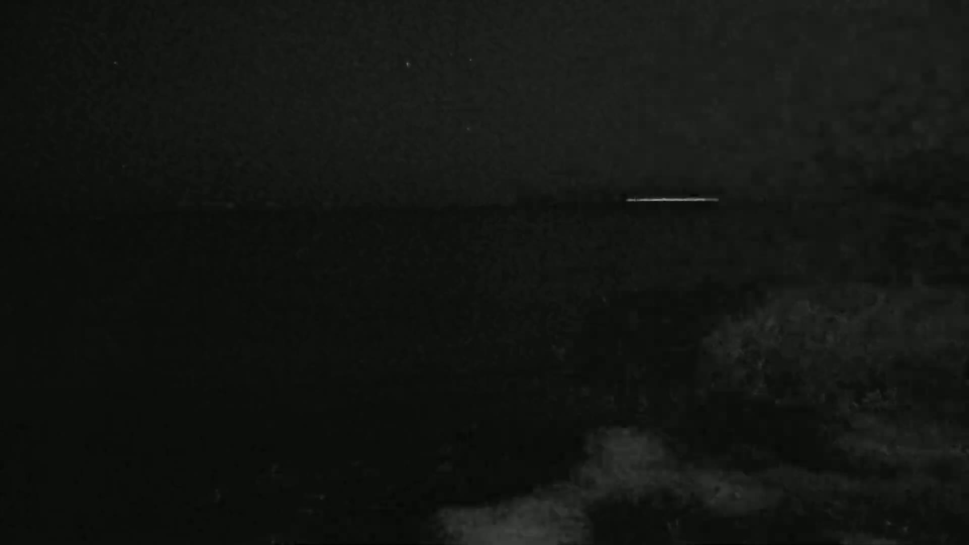 A dark, nighttime landscape shows a faint, horizontal light source in the distance over a mostly obscured body of water.