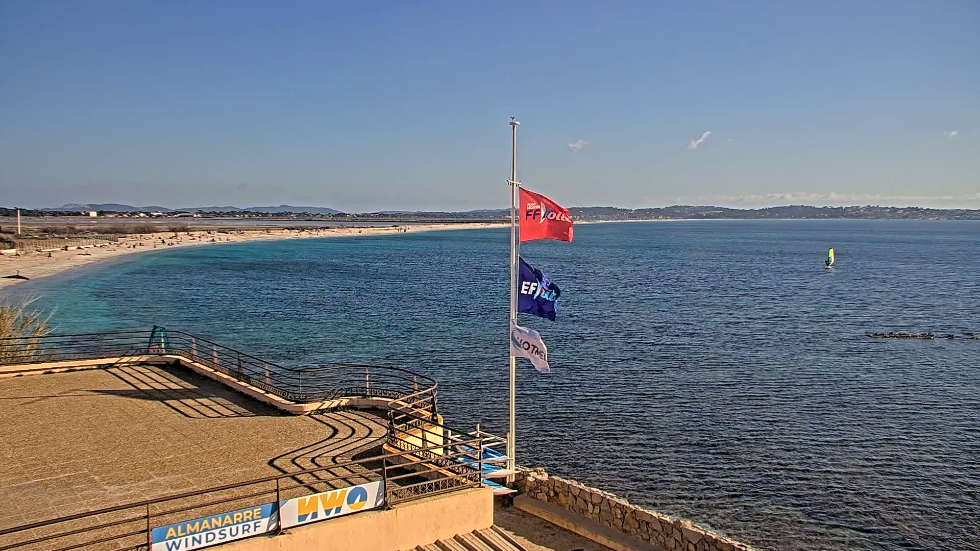 A sunny day overlooking a calm, turquoise ocean with a sandy beach and a windsurfer in the distance, from an elevated vantage point with a flagpole displaying several flags.