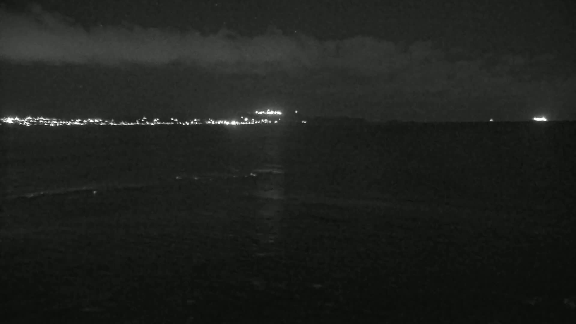 A dark, nighttime coastal scene shows a distant city skyline illuminated by lights under a partly cloudy sky.