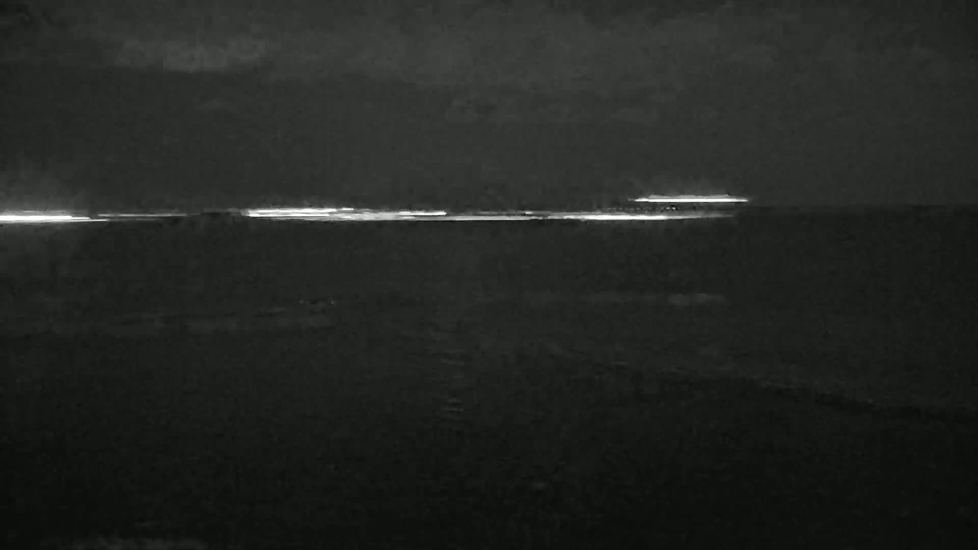 At night, a thin line of light from ships is visible on a dark body of water under a cloudy sky.