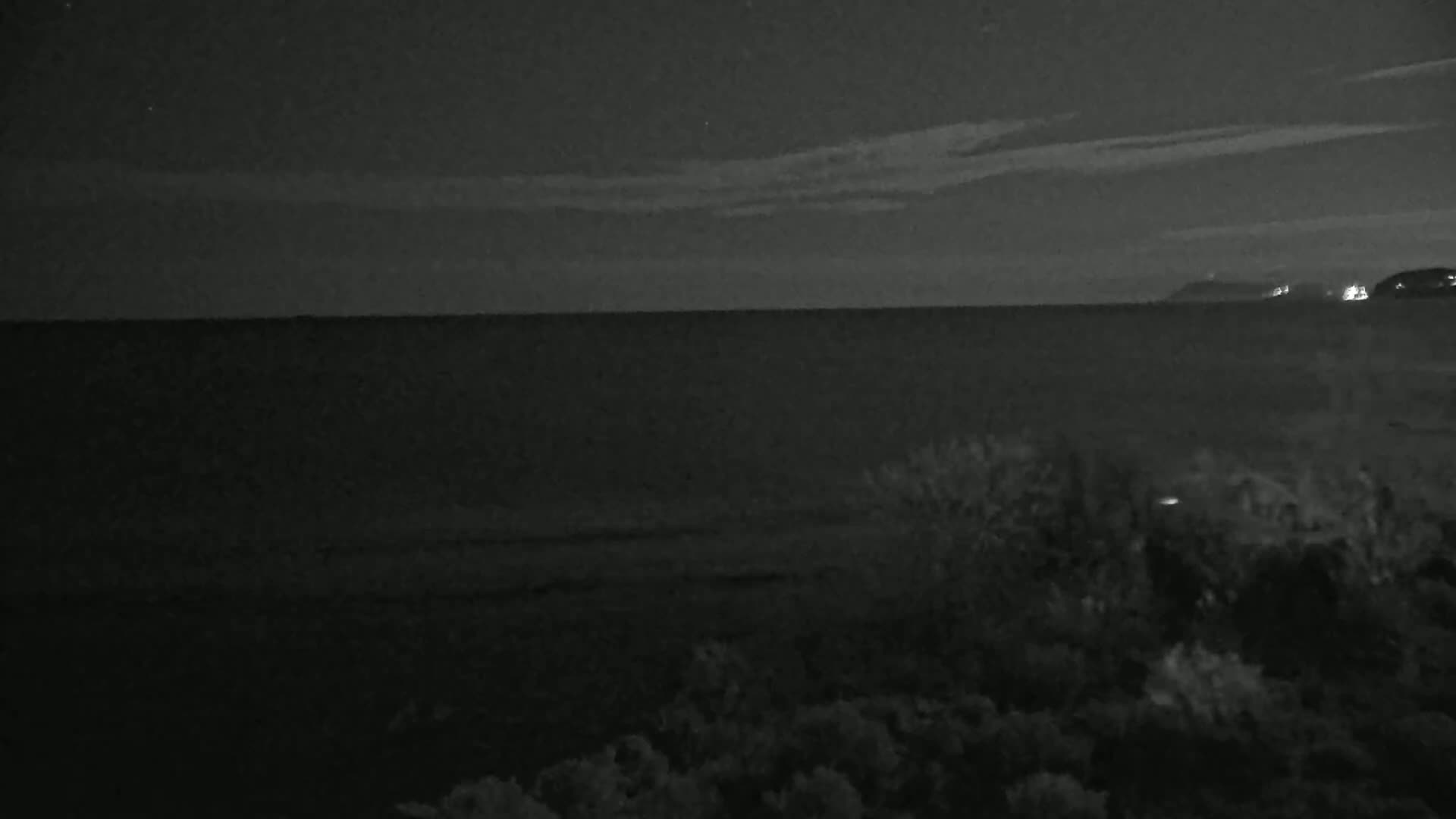 A dark, nighttime view of a calm sea under a mostly clear sky with a distant, faintly lit shoreline.