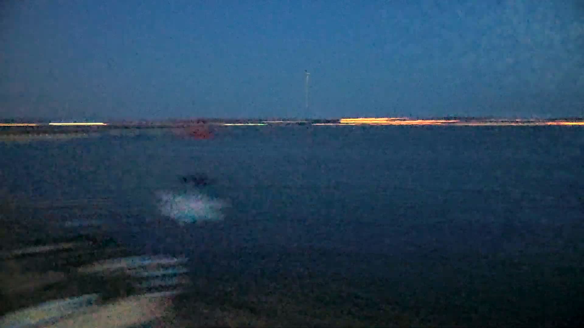 A dark body of water is shown at dusk, with a distant shoreline illuminated by faint orange lights.