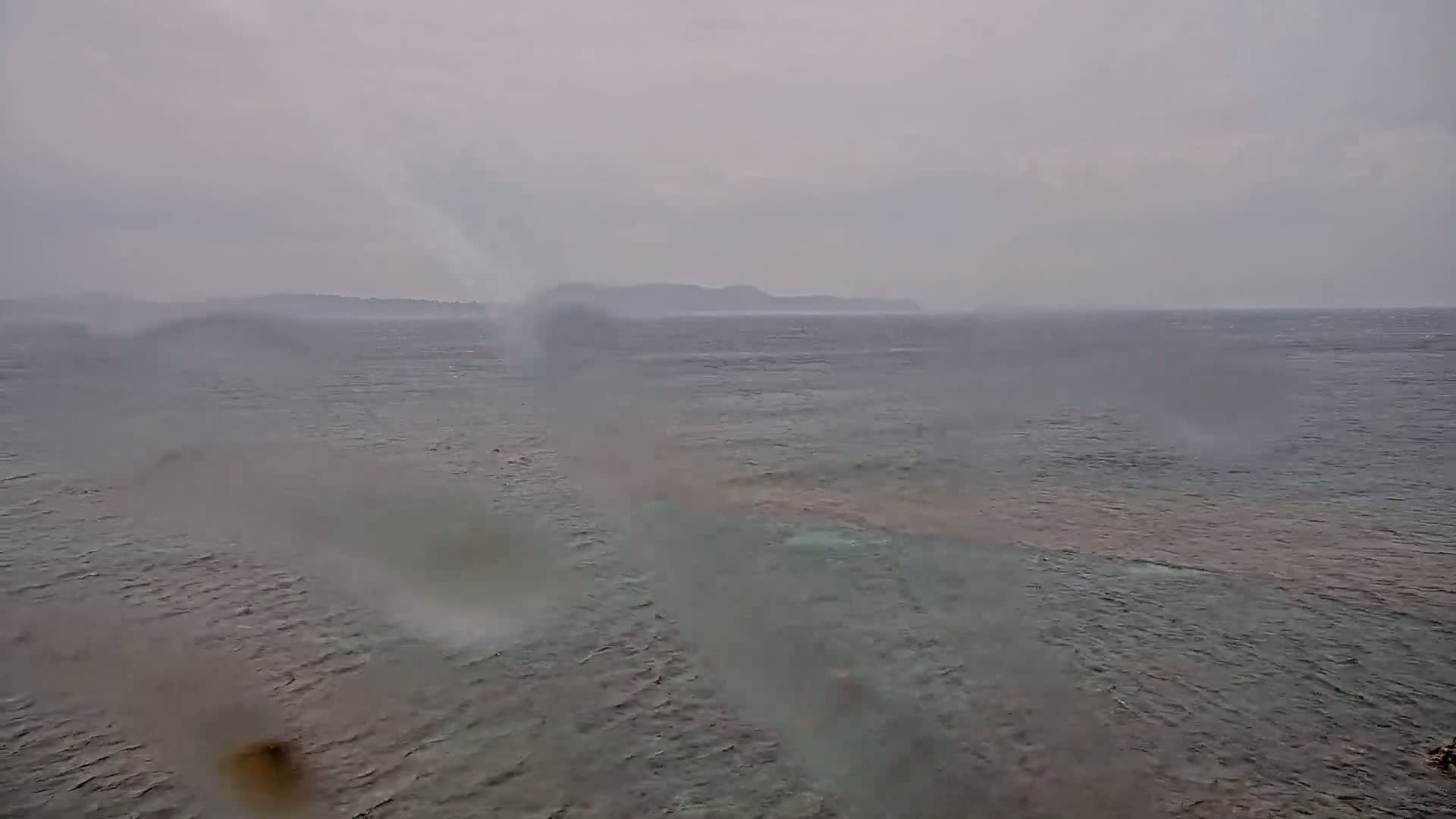 A view of a choppy sea with a distant shoreline under an overcast, rainy sky.