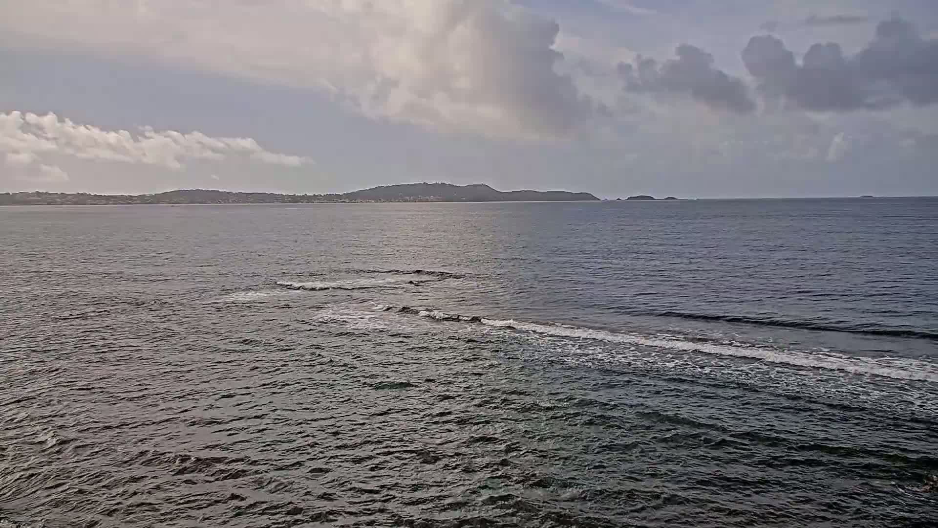 A partly cloudy day shows a wide expanse of dark-blue ocean with small waves lapping near the shore and a distant landmass under a mostly cloudy sky.
