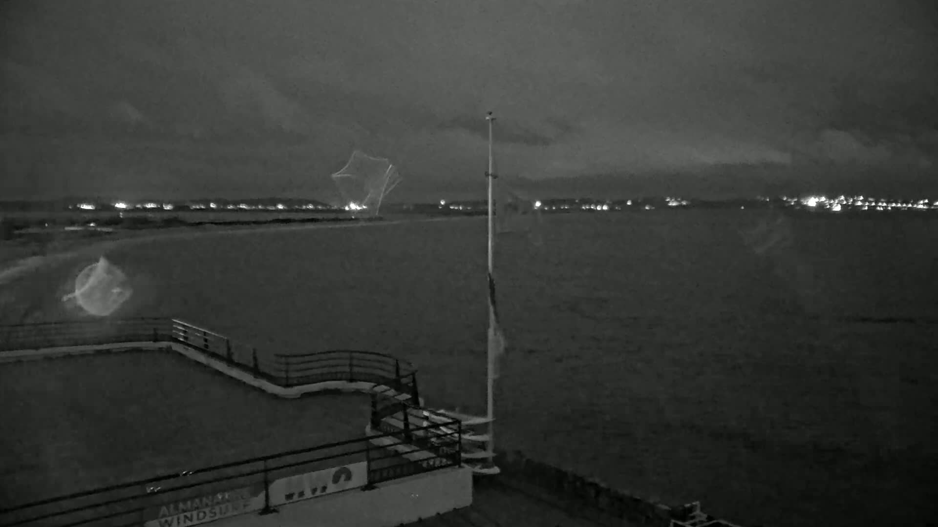 A nighttime view from an elevated position shows a dark body of water with a distant, faintly lit shoreline under a cloudy sky.