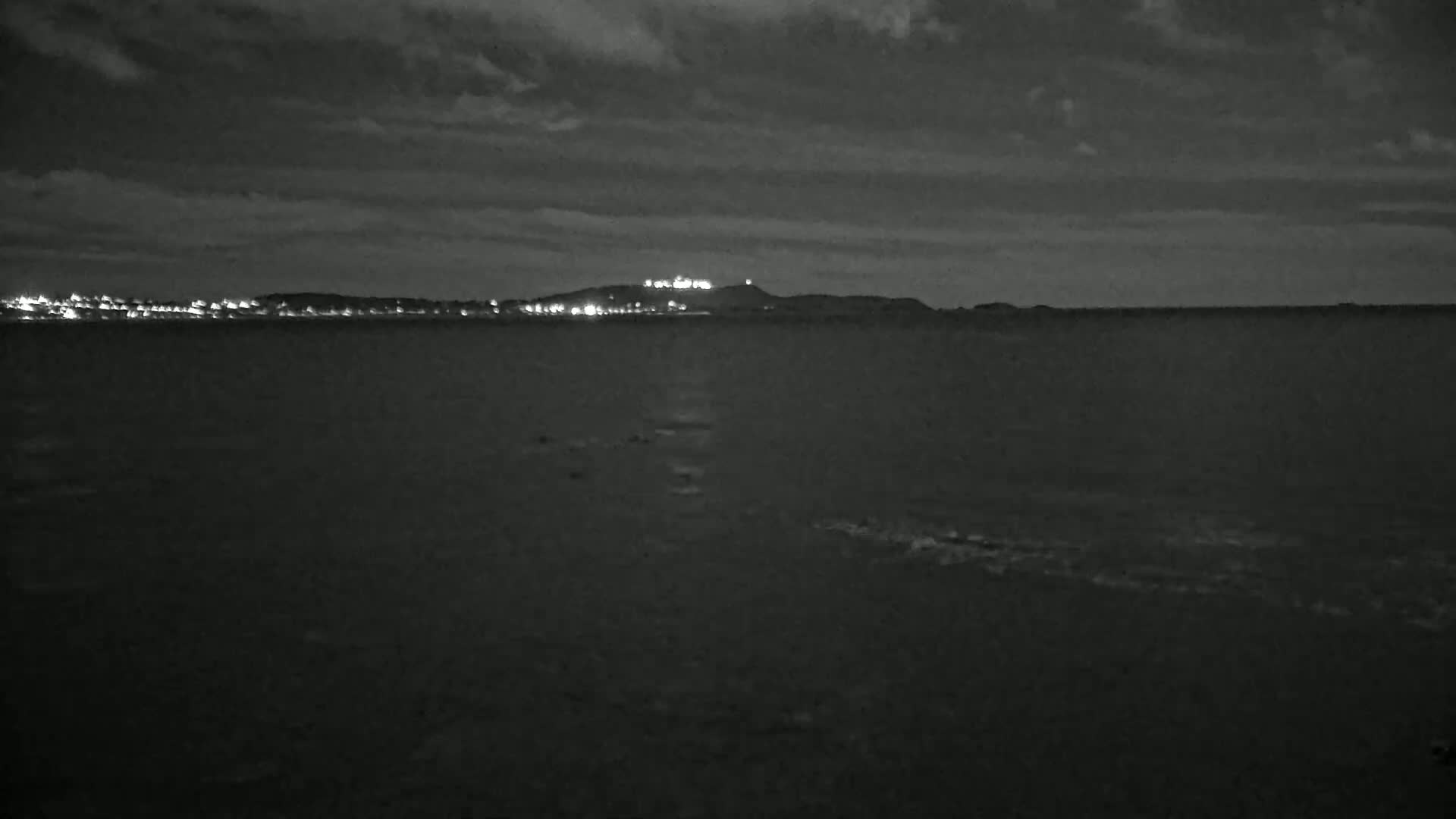 A dark, nighttime view of a calm body of water with a distant, dimly lit shoreline and hill under a cloudy sky.