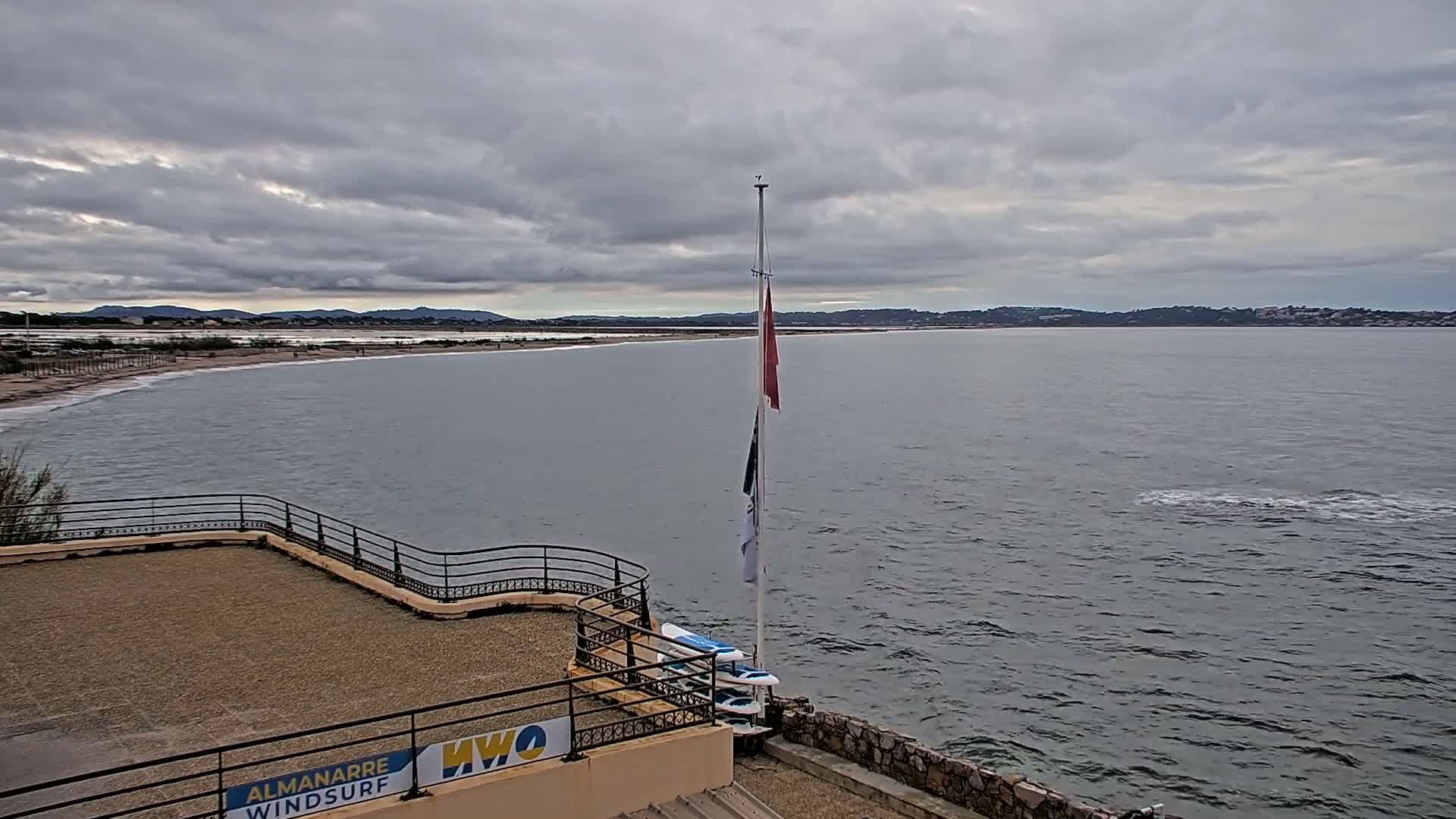 A calm, grey day shows a view of a body of water, a sandy beach, distant hills, and a flagpole with several flags on an elevated seaside structure.