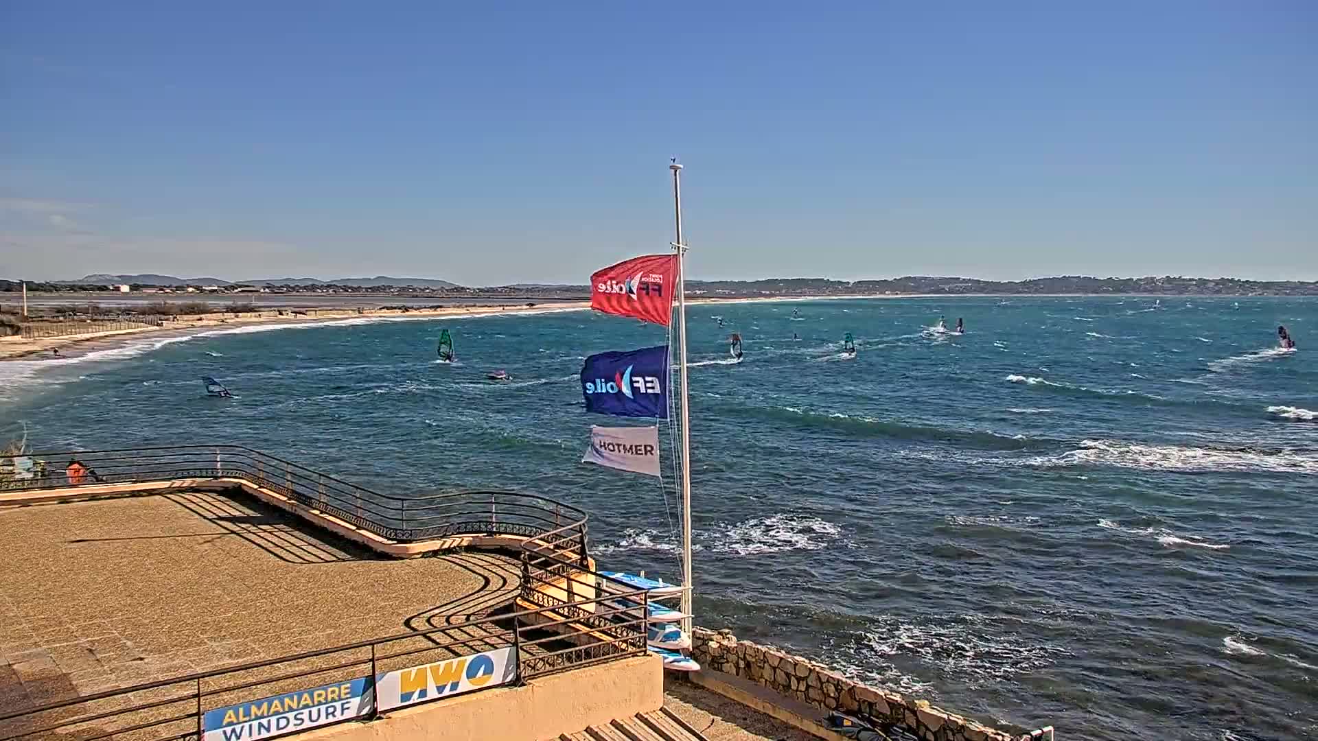 A mostly sunny day shows a beach with several windsurfers on a wavy ocean, viewed from an elevated vantage point with a flagpole and concrete structures nearby.